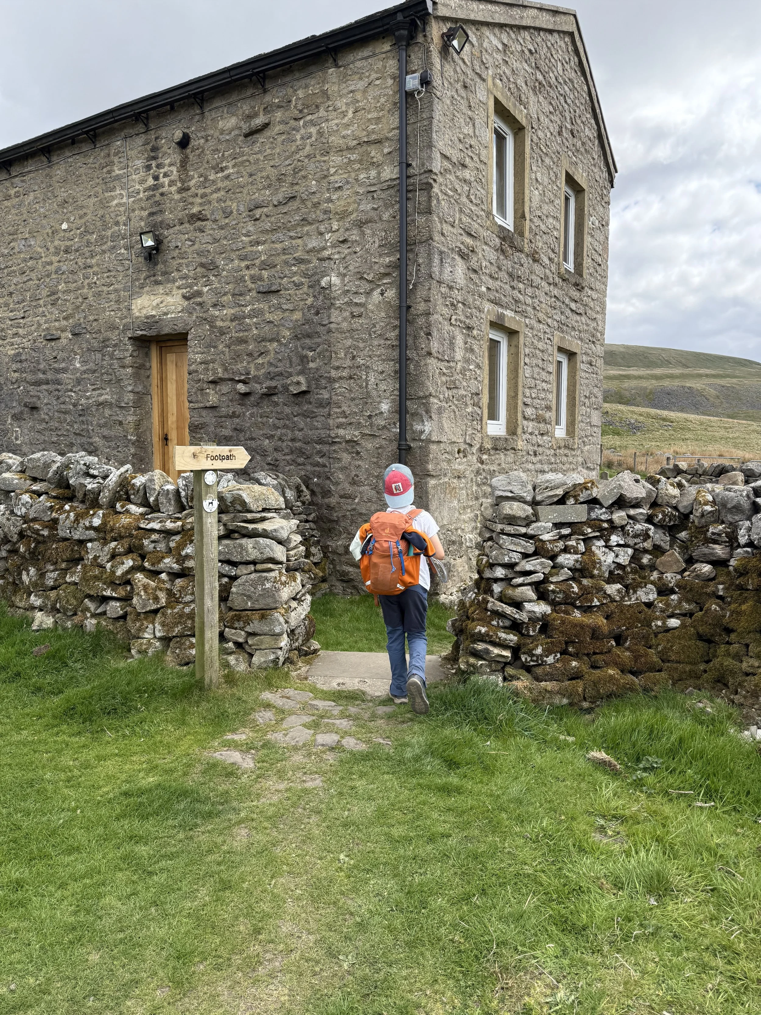 Child walking past stone built building through a dry stone wall with a footpath sign wearing a backwards cap and orange backpack