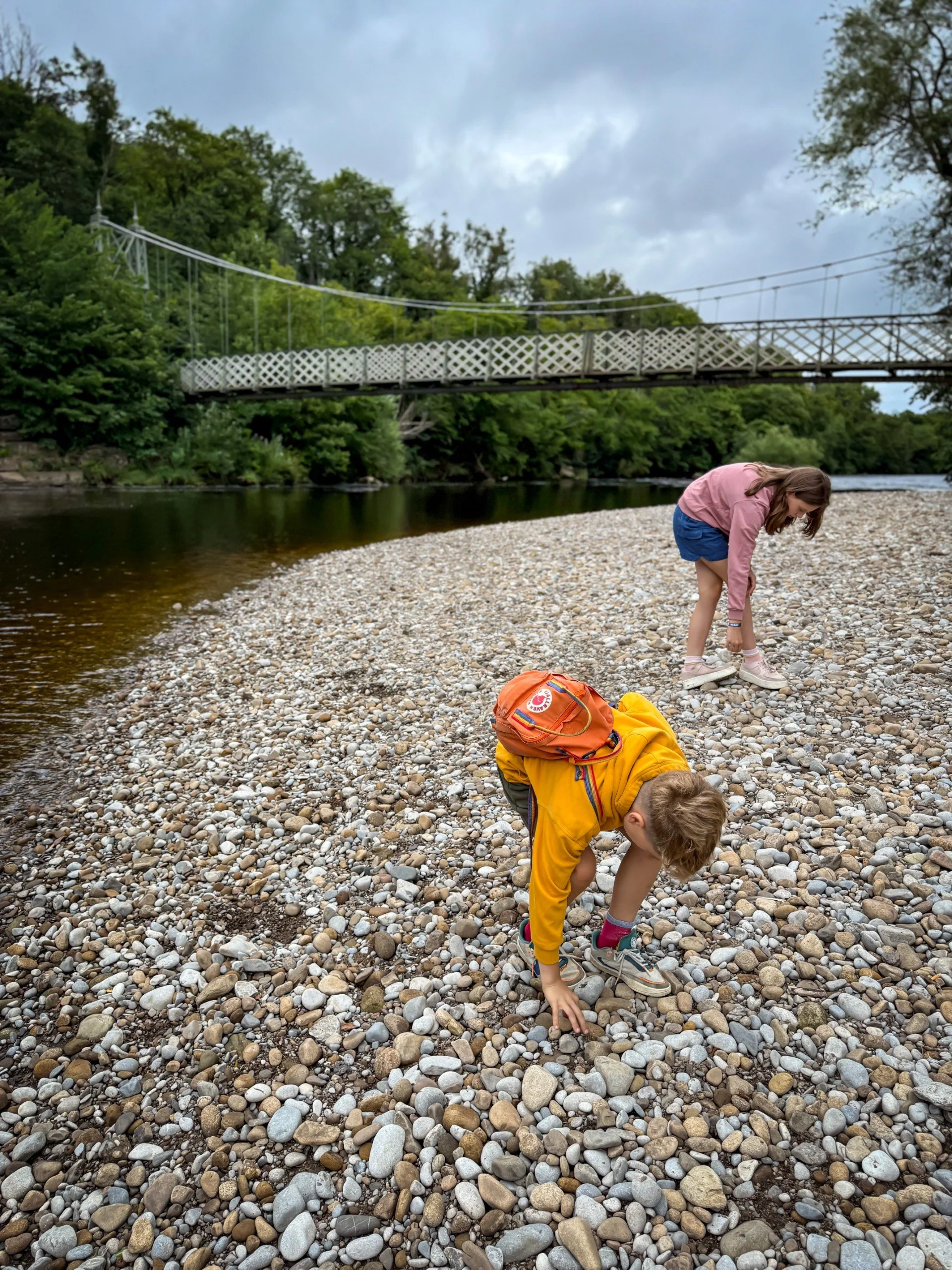 Two children bent over picking up stones to throw into the river they are next to with a bridge behind