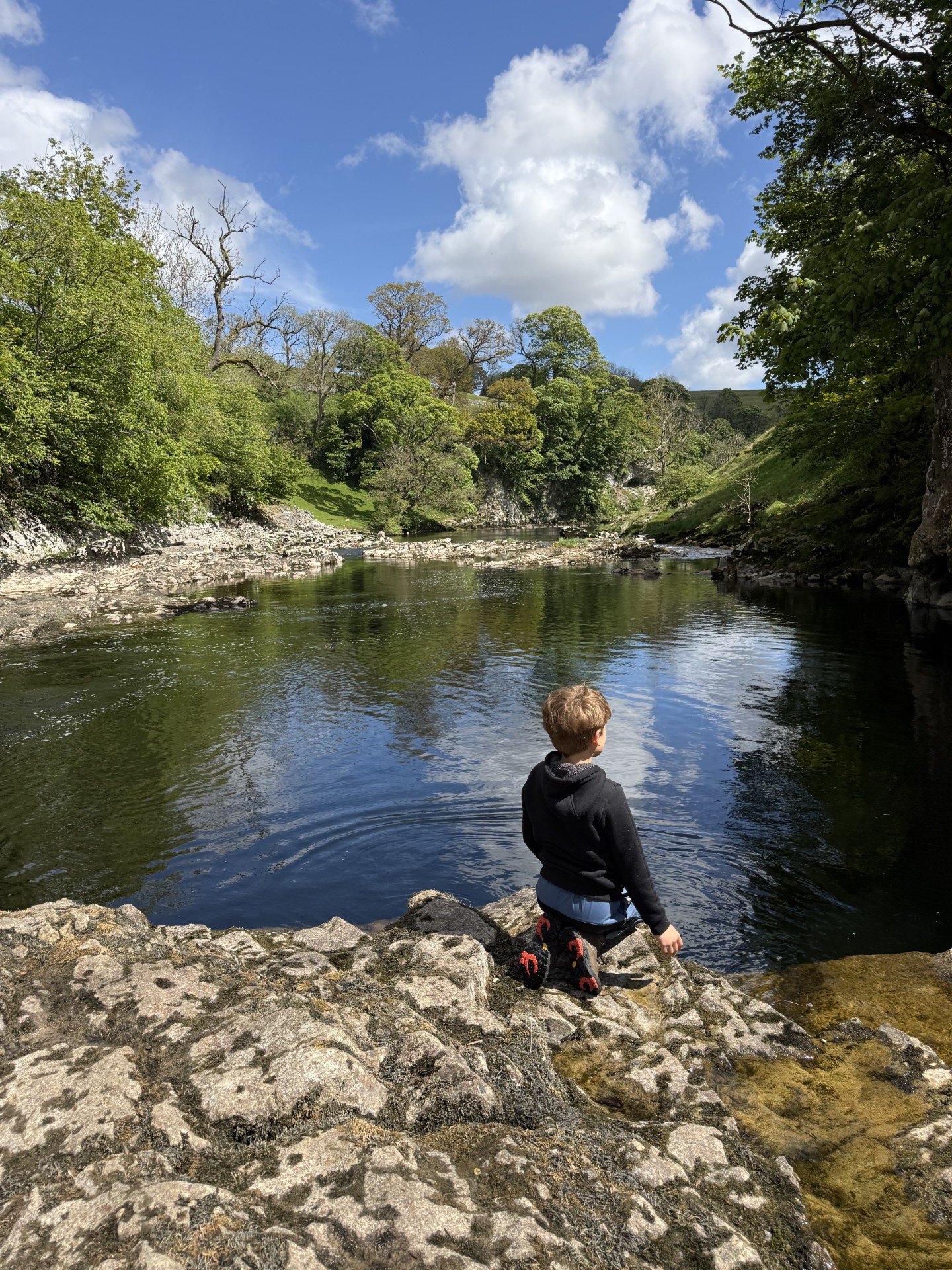 Child kneeling down by a river with rocky cliff sides and green trees