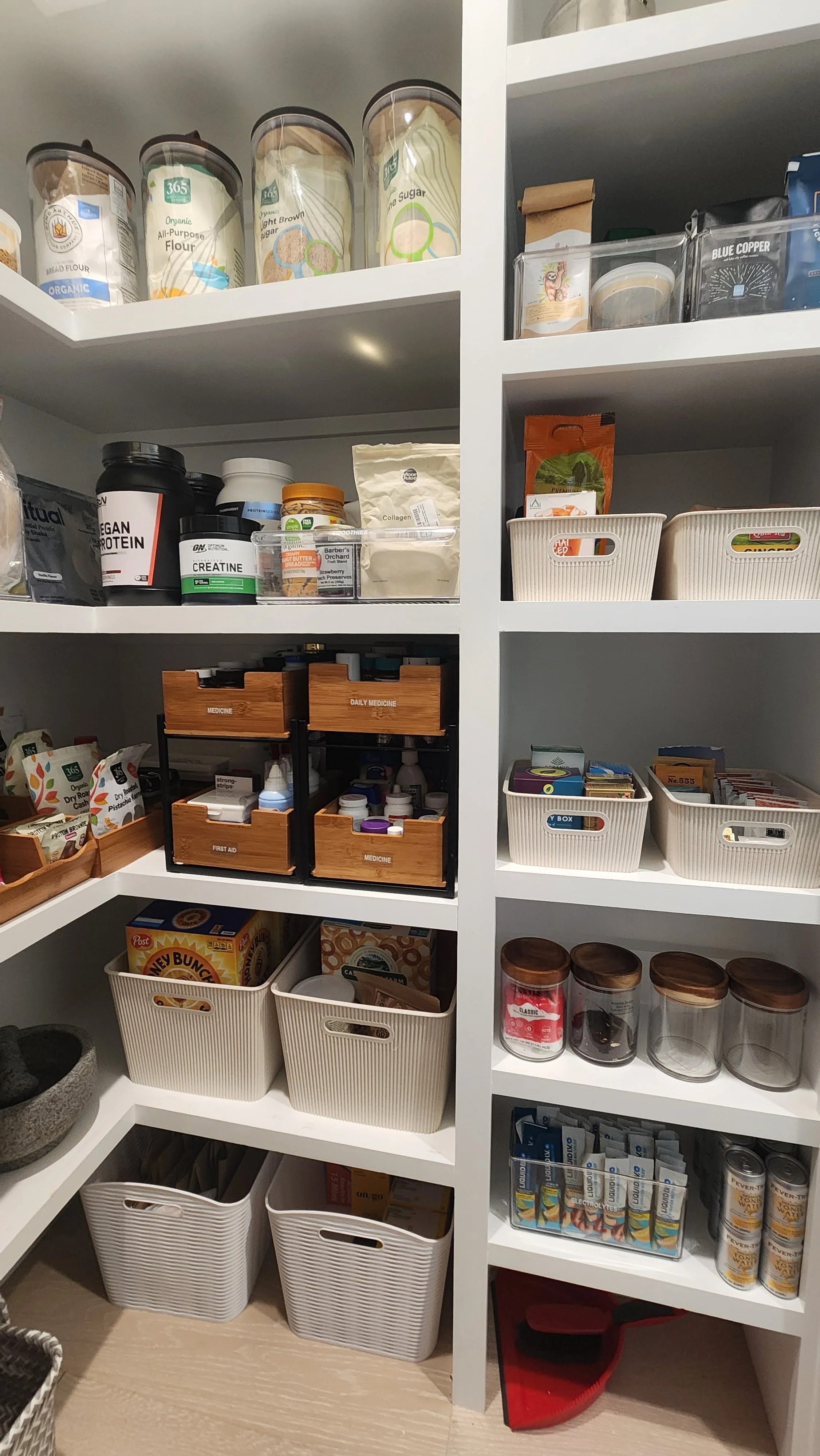 A well-organized pantry with labeled containers, shelves holding various food items, supplements, medication boxes, and snacks in baskets.
