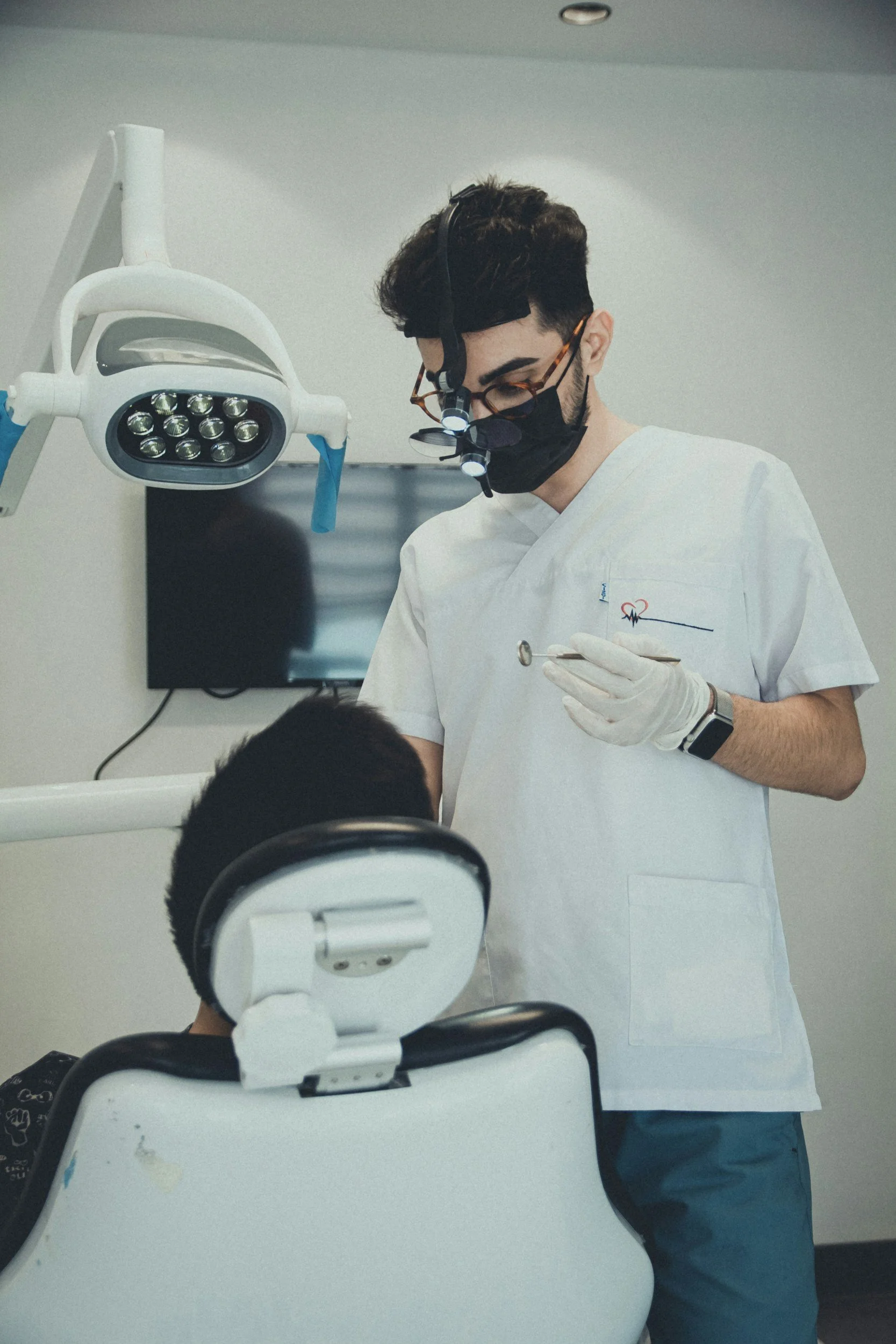 A male dentist wearing glasses, a face mask, and magnifying loupes with lights, is examining a patient in a dental clinic. The patient is seated in a dental chair with the back of their head visible, and the dentist is holding a dental tool.