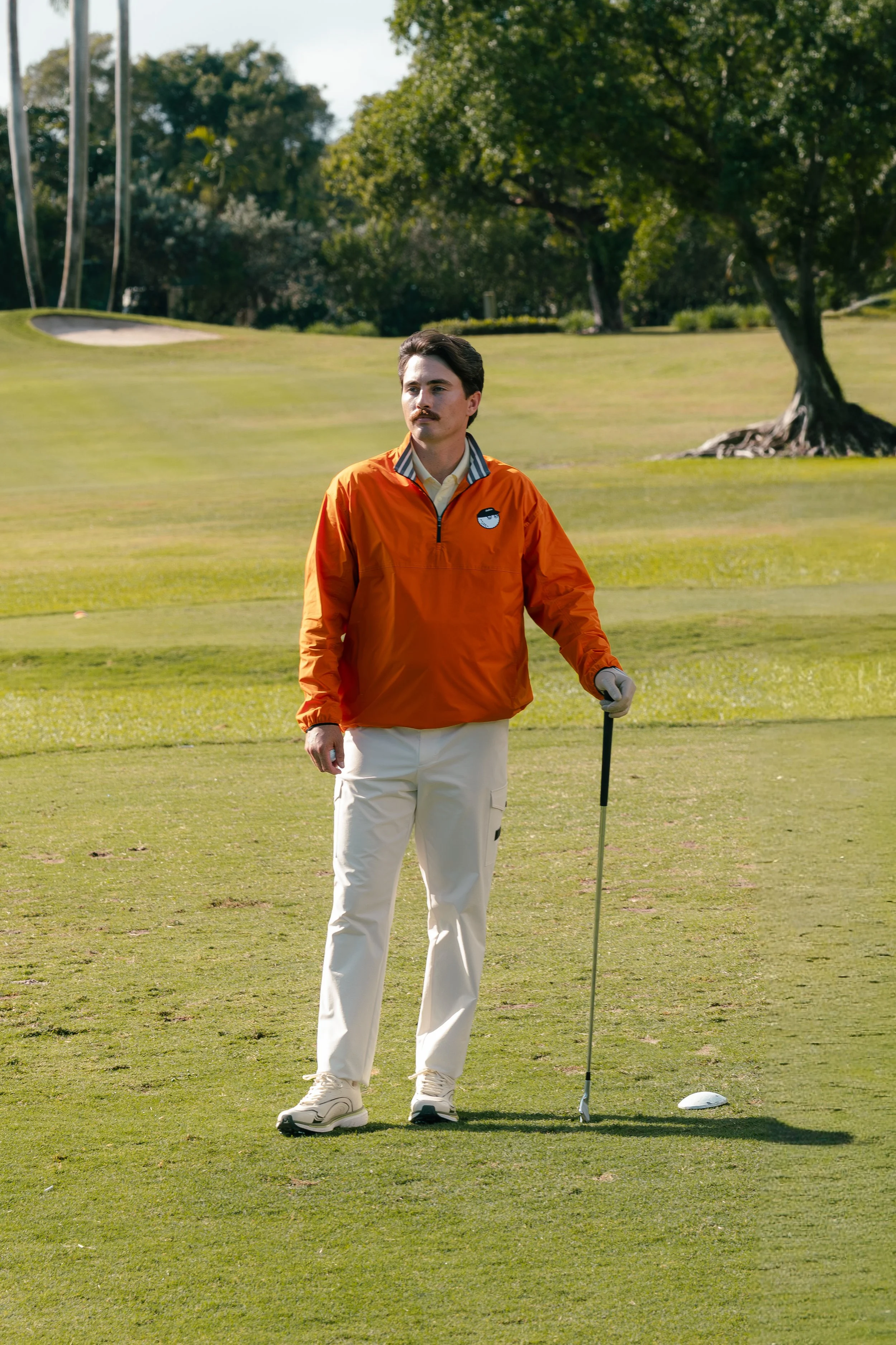 A man with a mustache is standing on a golf course, holding a golf club, next to a golf ball teeing off.