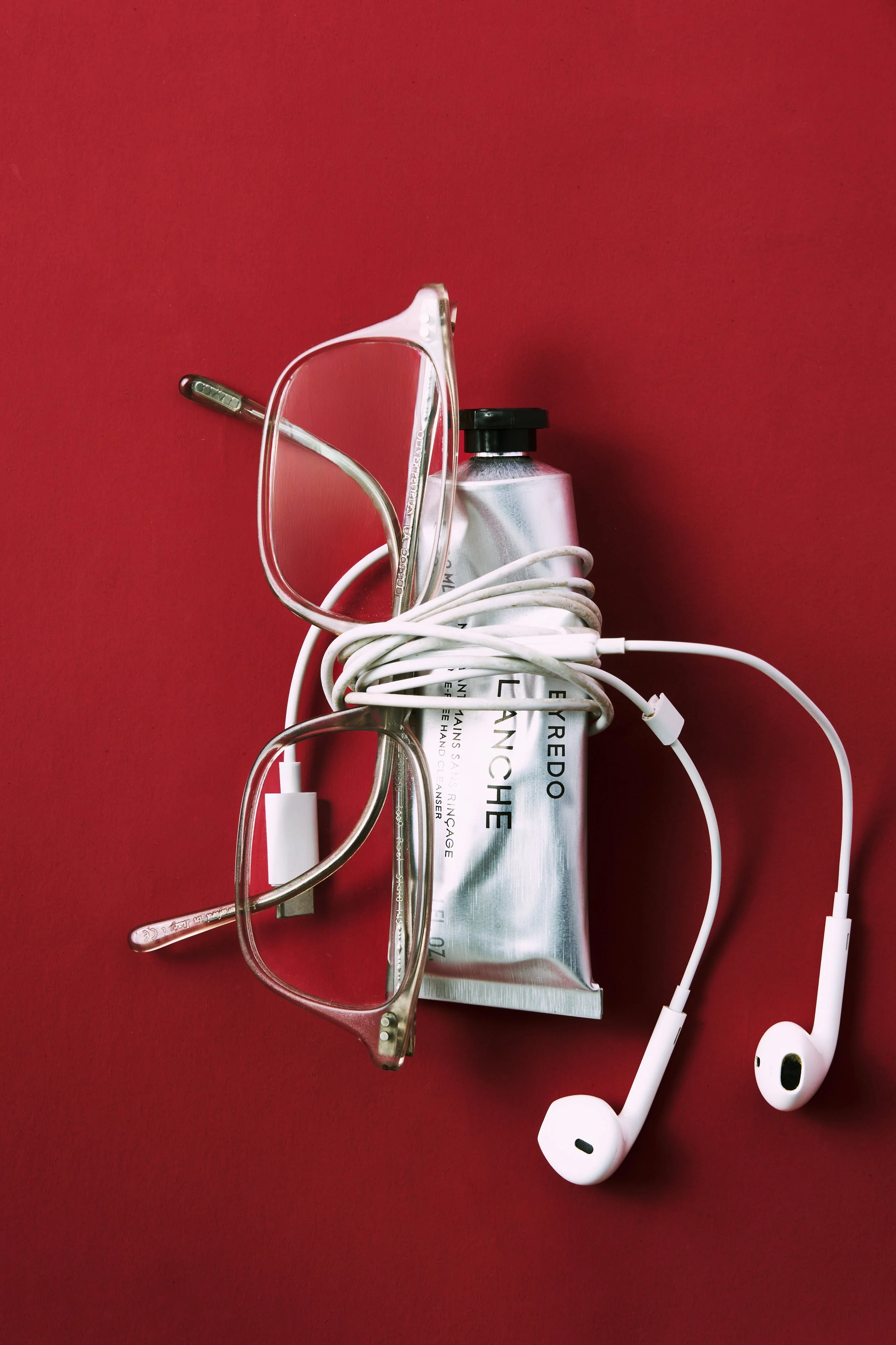 Eyeglasses, a tube of cream, and a pair of white earphones on a red surface. Byredo