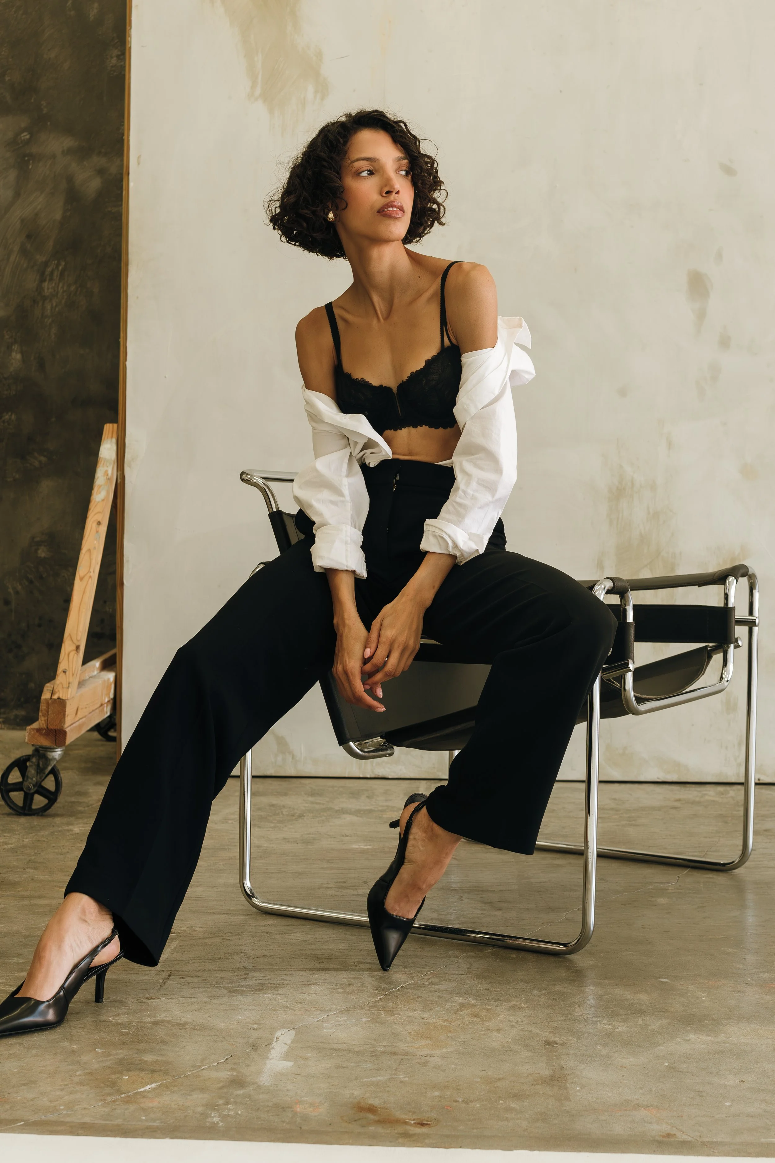 Test Shoot. A woman with curly hair in black heels, black pants, a black lace bra, and an unbuttoned white shirt sitting on a chair in an industrial setting.