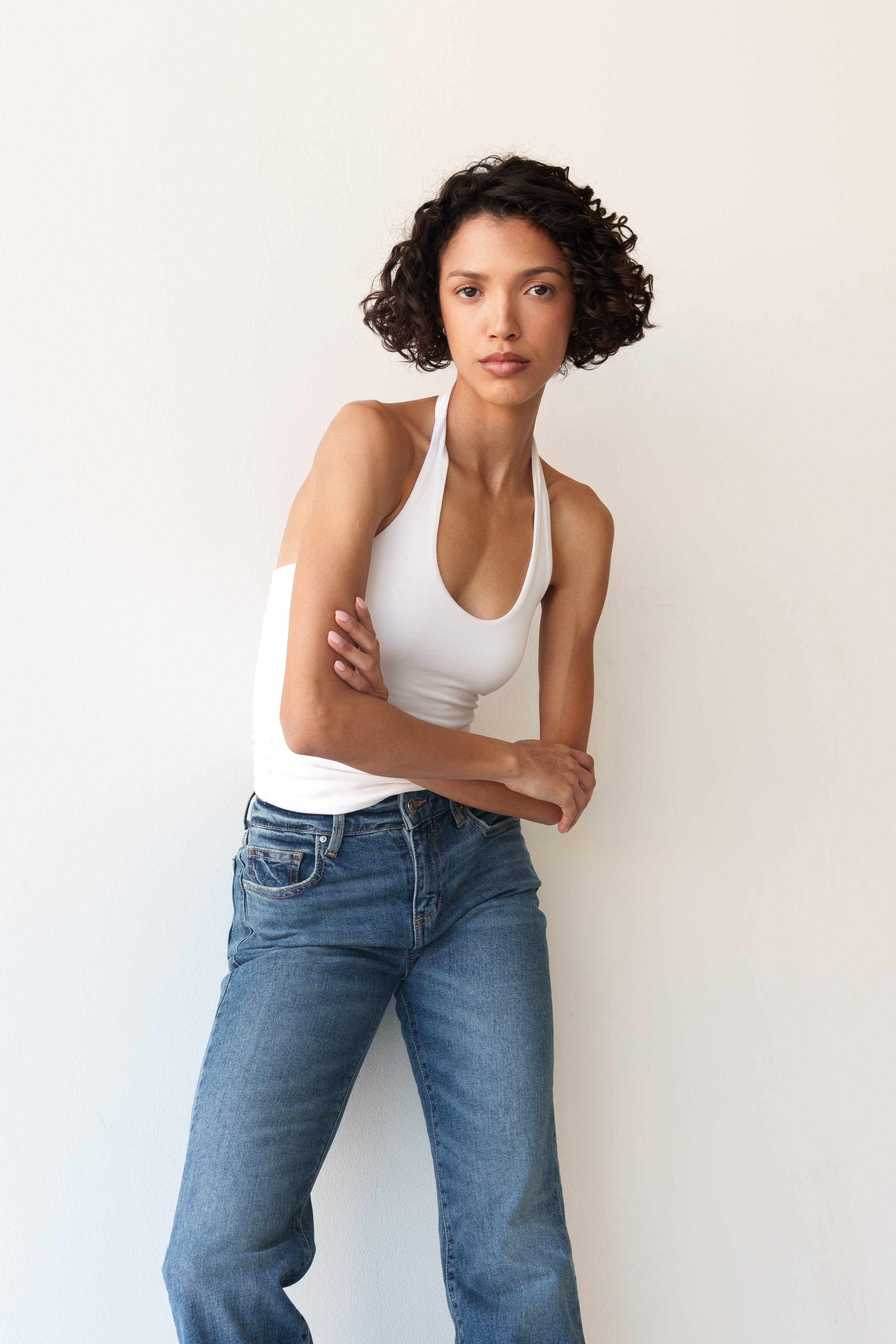 Test Shoot A young woman with curly dark hair, wearing a white tank top and blue jeans, standing against a plain white wall, looking directly at the camera.