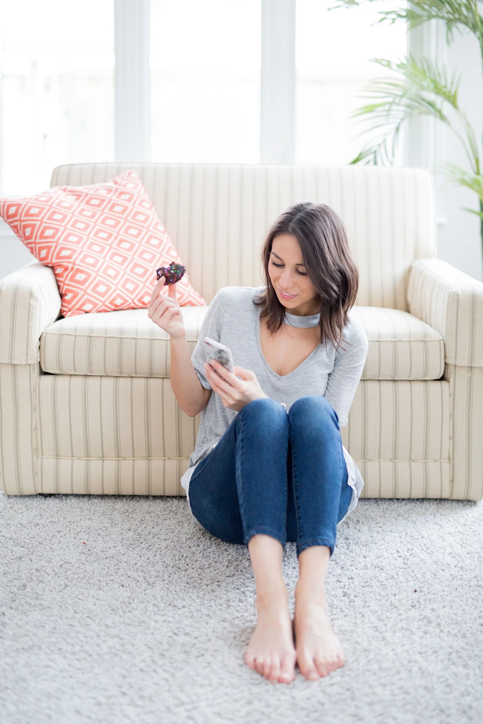 A woman sitting on a carpeted floor in front of a sofa, holding a donut with colorful sprinkles in her right hand and a phone in her left hand while looking at it. She is wearing a gray t-shirt and blue jeans, with her legs crossed at the ankles. The sofa behind her has a pink and white geometric patterned pillow. There is a large window in the background with white curtains, and a tall green plant to the right.