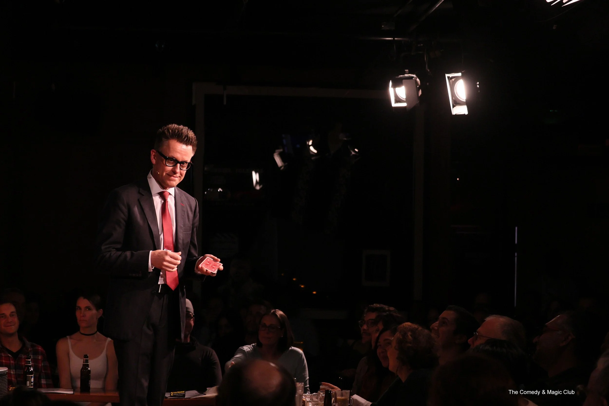 Arthur Trace on stage at The Comedy & Magic Club. He is in a suit and addressing the audience.