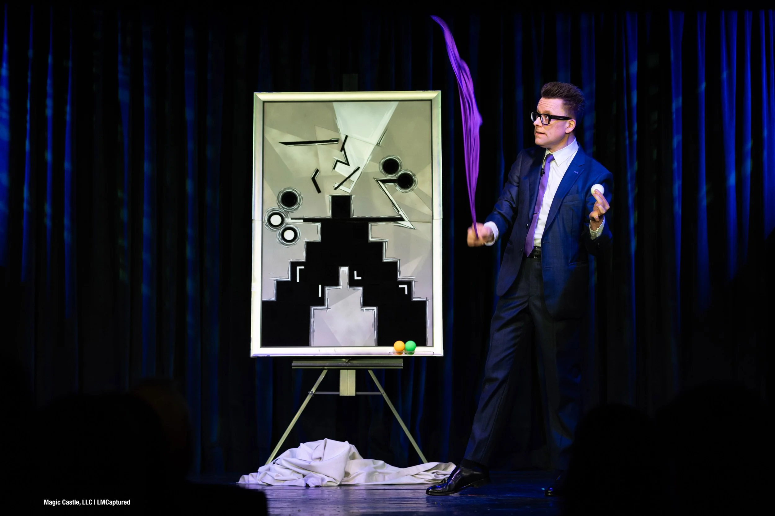 Arthur Trace on stage at The Magic Castle. He holds a ball in one hand and flicks a purple silk with the other. An abstract painting is behind him.