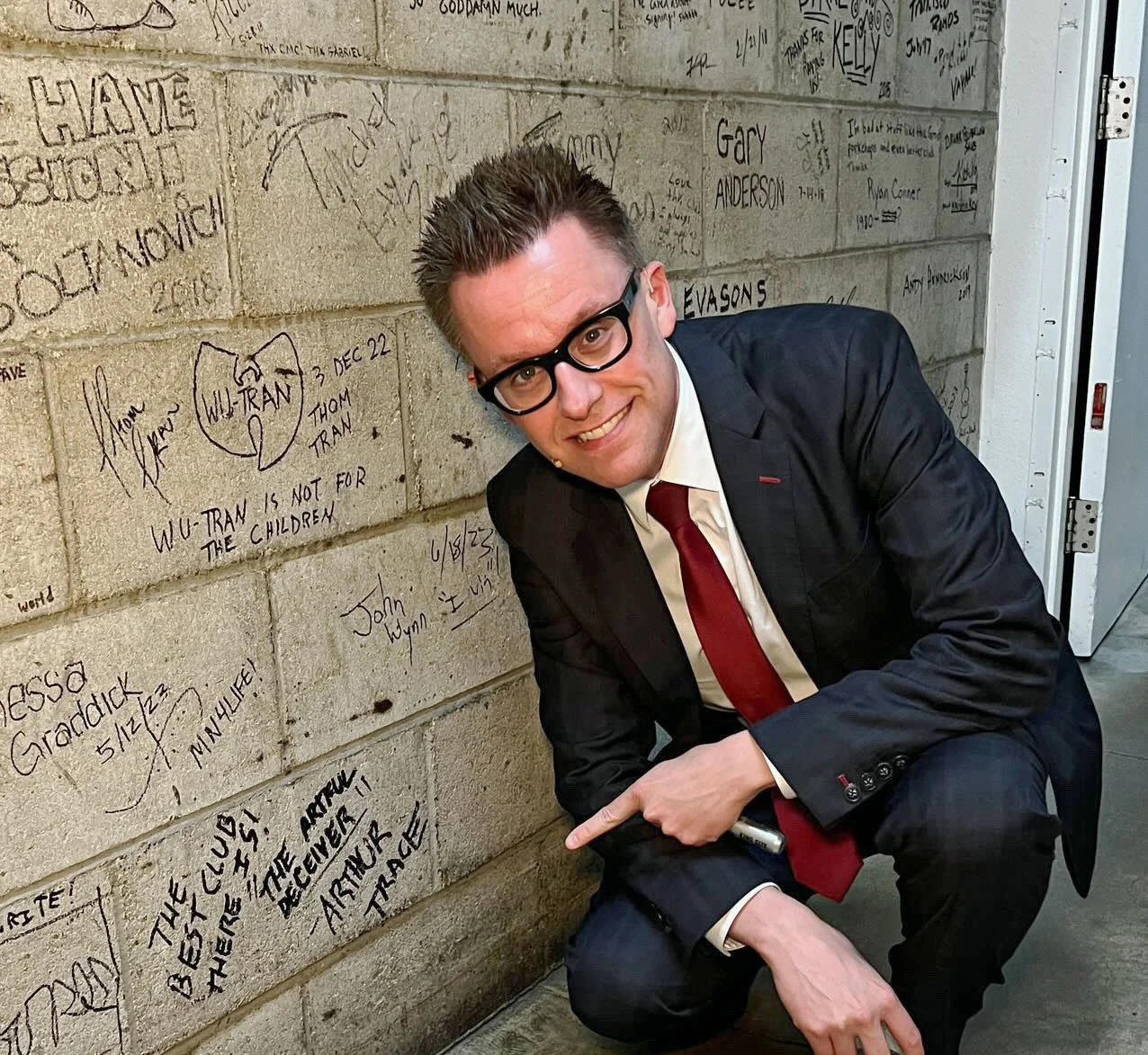 Arthur Trace backstage at The Comedy & Magic Club. He is pointing to a brick wall where he signed his name.
