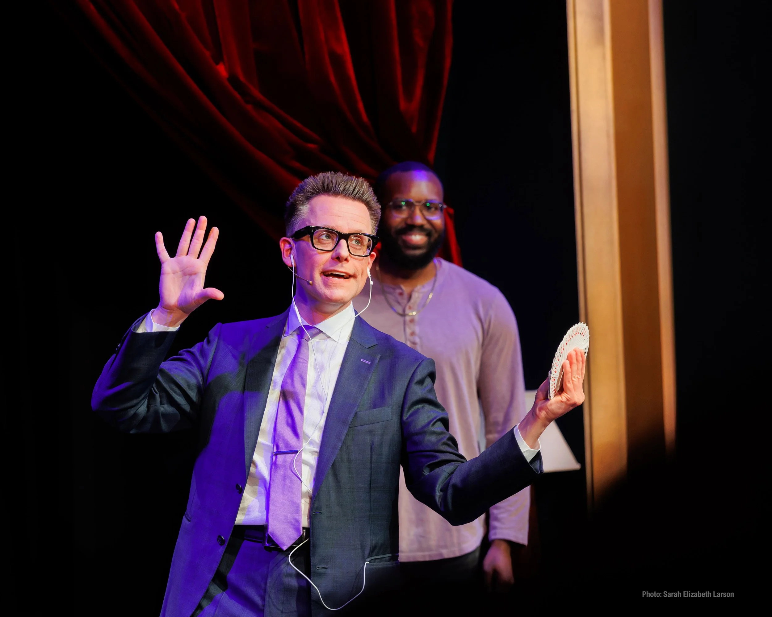 Arthur Trace performs a card trick on stage at The Chicago Magic Lounge. Arthur is holding a fan of playing cards while a male volunteer looks on.