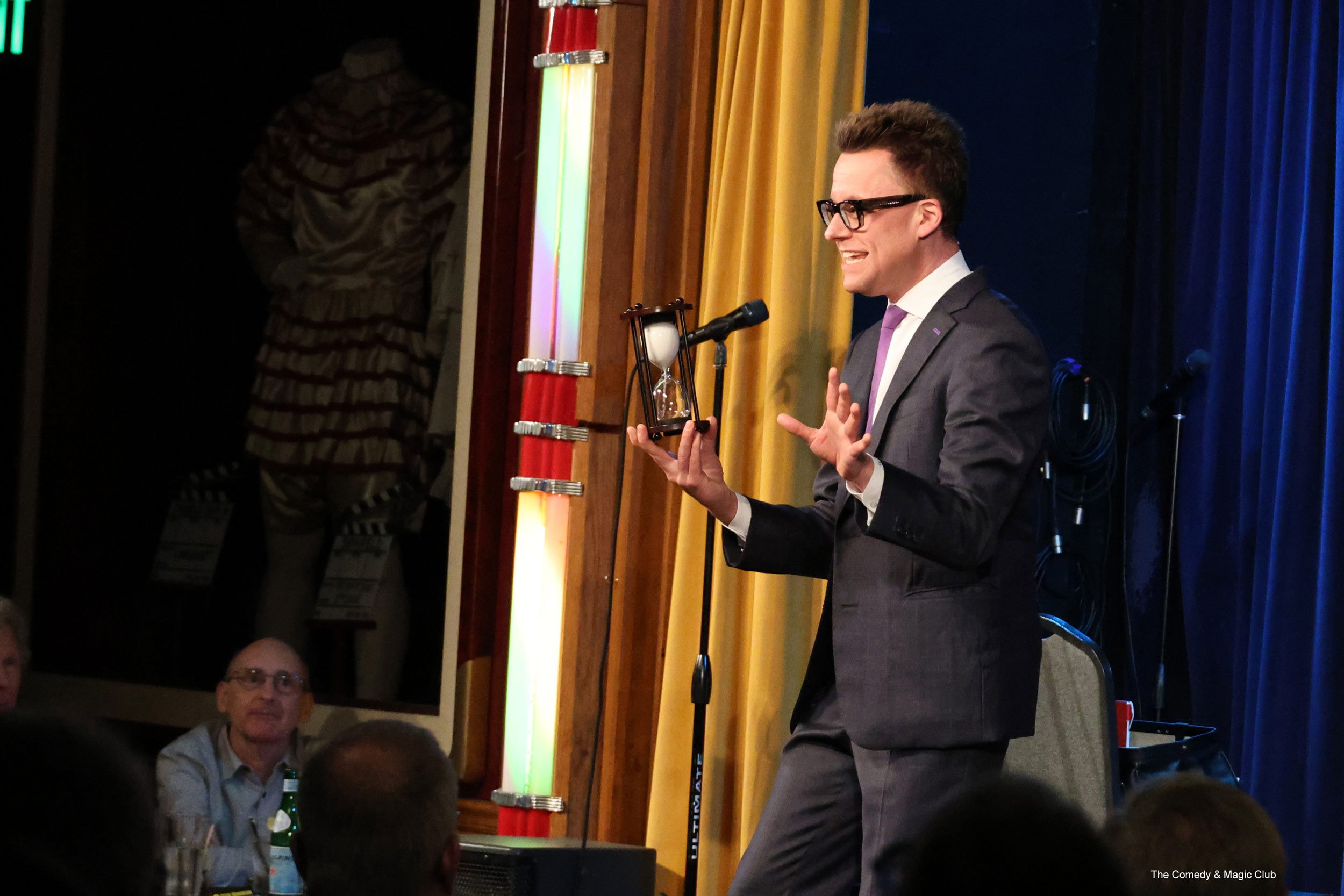 Arthur Trace on stage at The Comedy & Magic Club. He is holding an hourglass and addressing the audience.