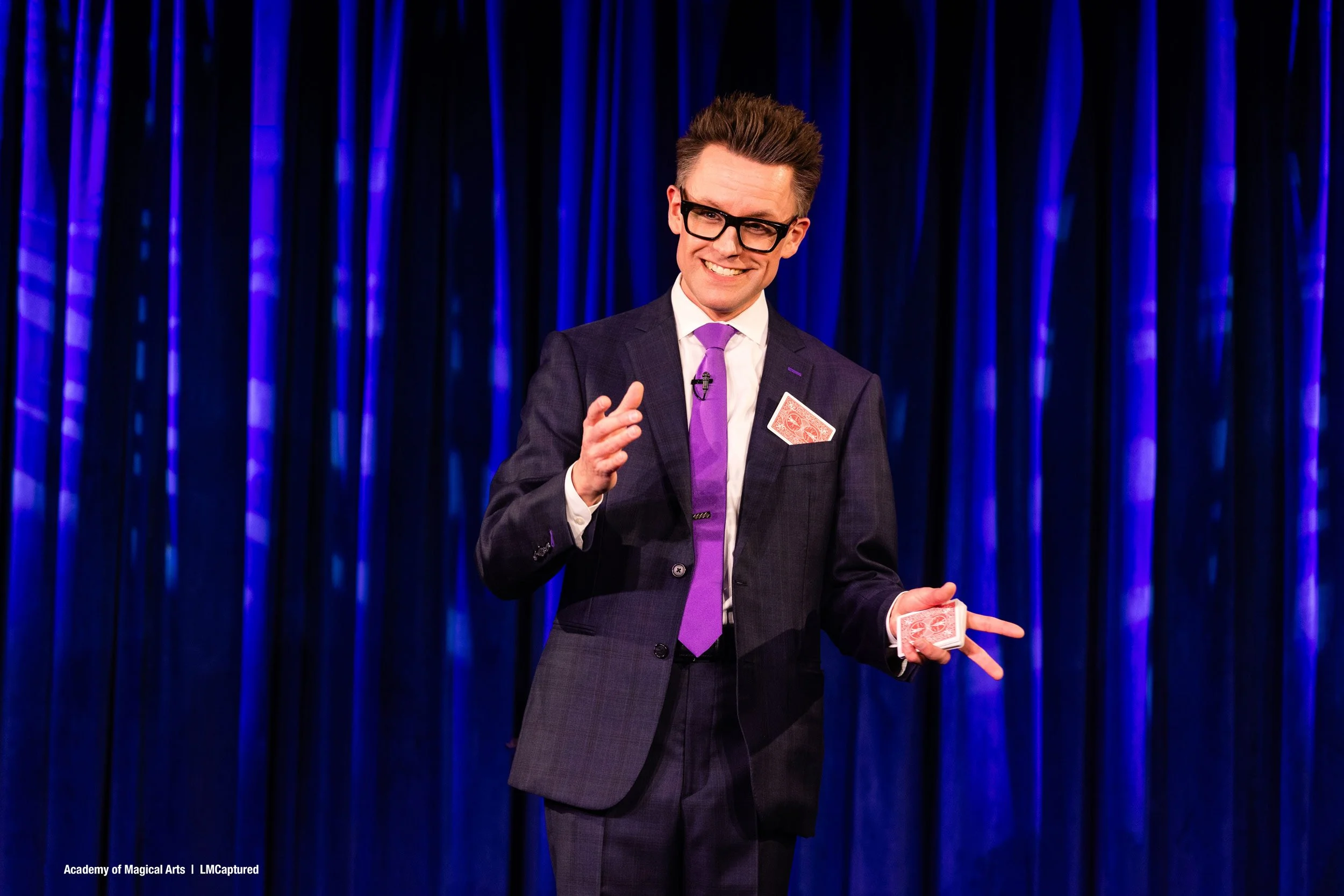 Arthur Trace stands smiling on stage. He has a deck of playing cards in his left hand and one card sticking out of his pocket.