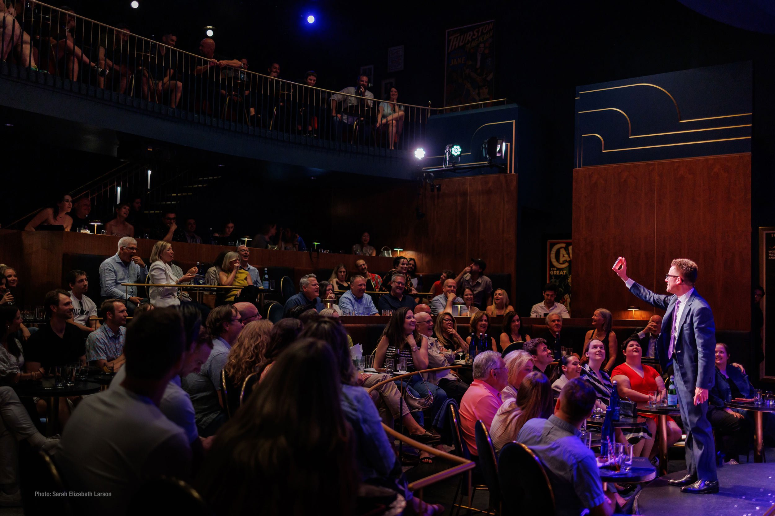 Arthur Trace performing a card trick on stage as a large audience looks on.