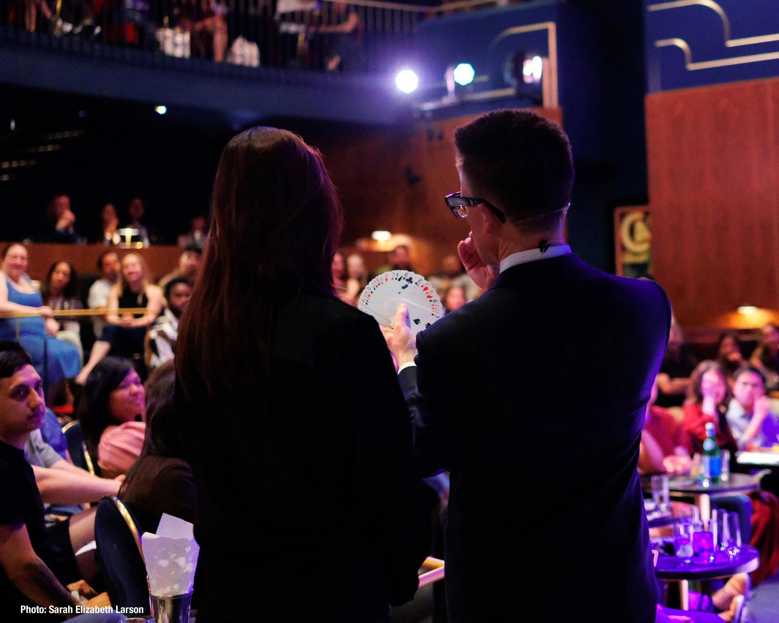 Arthur Trace, seen from behind, fanning a deck of cards for an audience member during a performance in a theater, with rows of seated guests watching on either side.