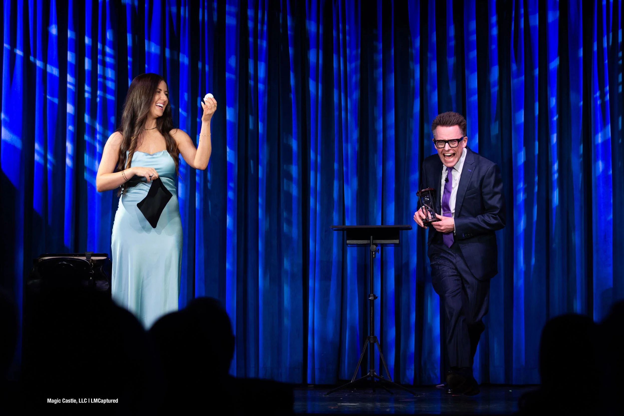 Arthur Trace and a female volunteer are pictured on stage. The volunteer is holding an egg and smiling. Arthur is holding an hourglass.