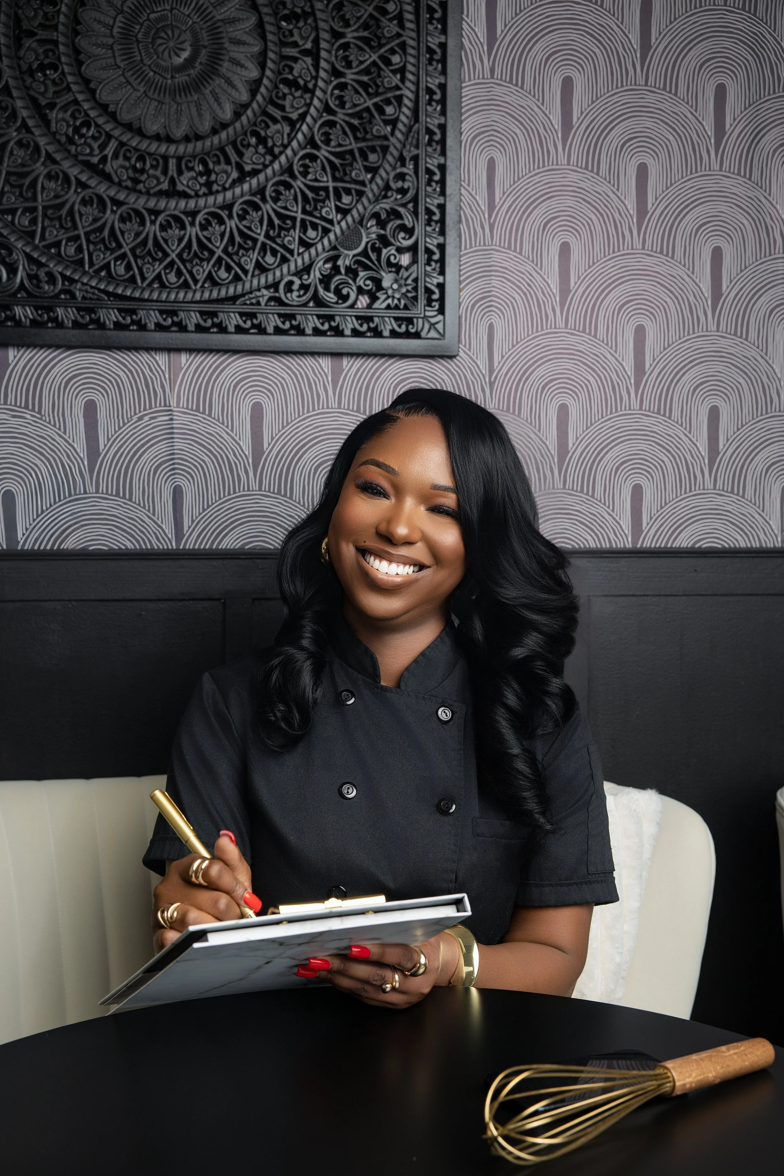 Smiling woman in black chef coat sitting at a black table with a gold whisk beside her, holding a notepad and pen.