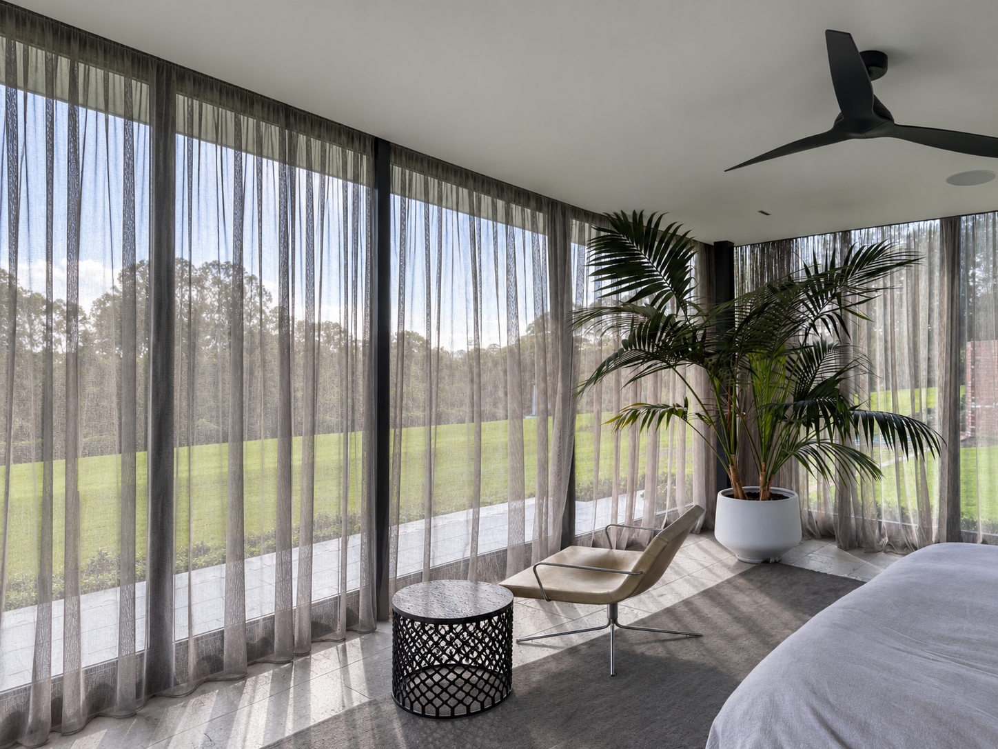 Living room with a large potted plant near glass windows with curtains, ceiling fan, lounge chair, and sofa, overlooking a green outdoor area.