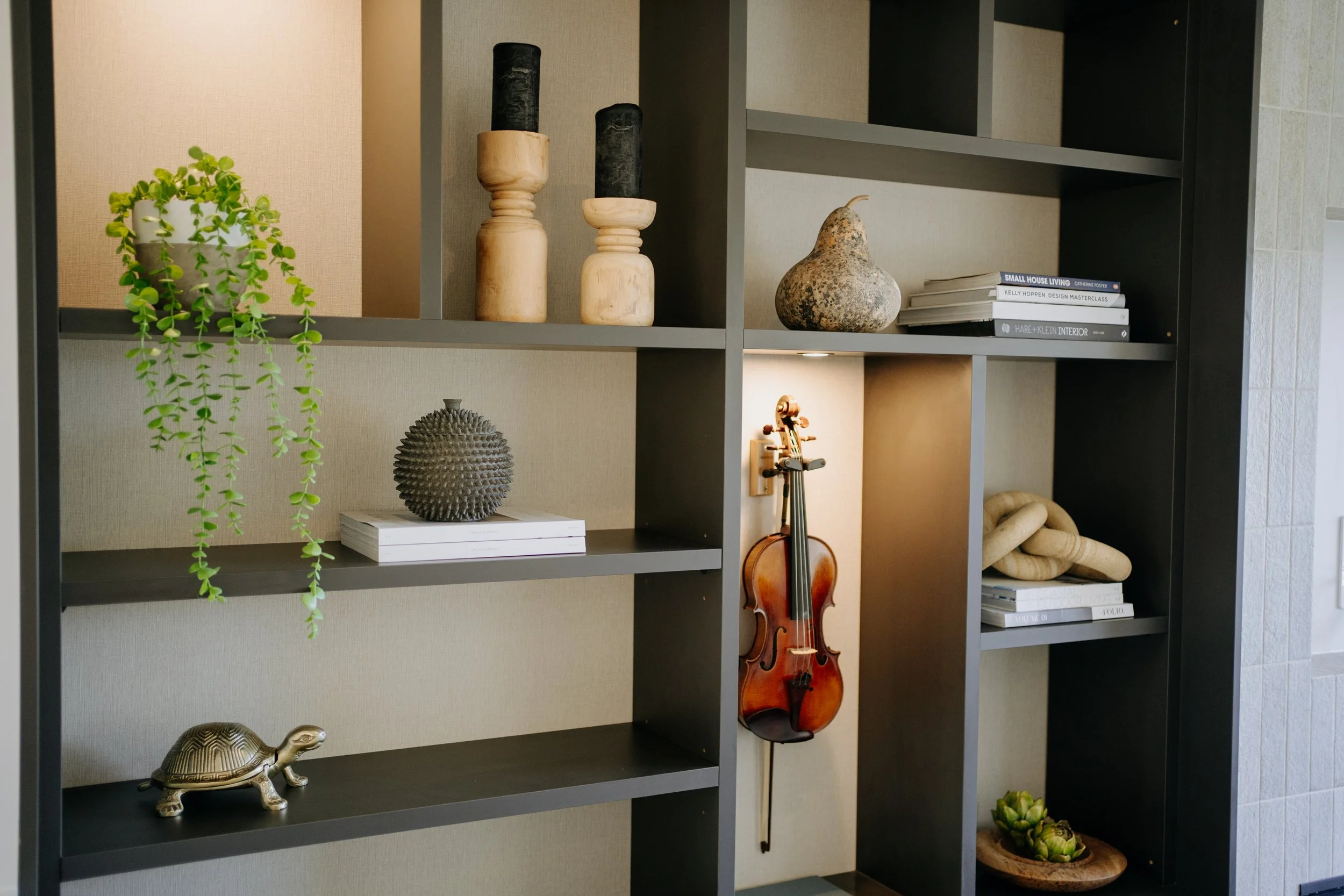 Bespoke cabinetry designed by McMahon Interior Designer for a North Shore Auckland Bungalow. Shelf with decorative items including a potted plant, candles, a stone ornament, books, a violin, and a turtle figurine.