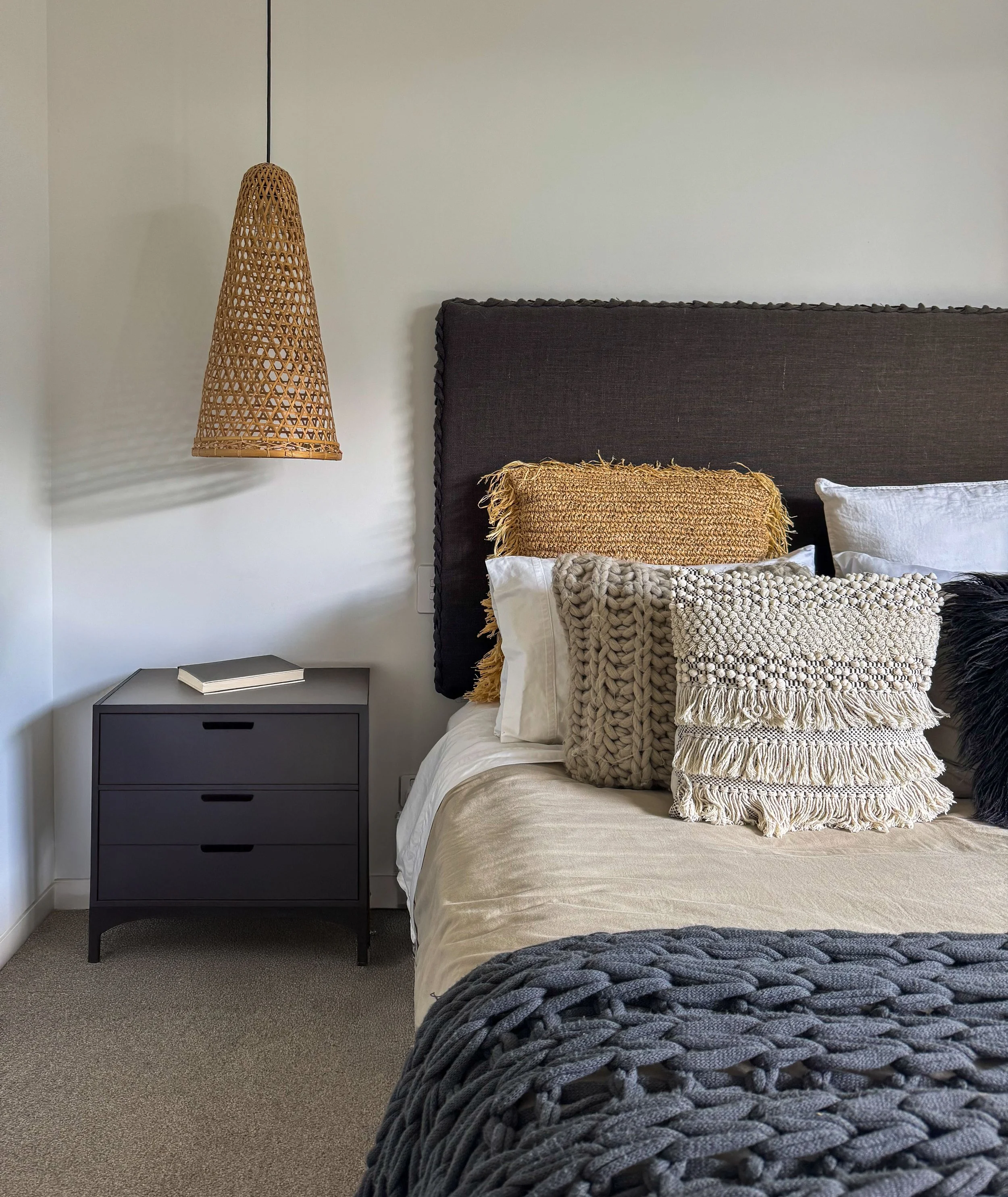 A bedroom with a dark gray headboard, beige bedding, multiple pillows including a chunky knit cream pillow, a white pillow, and a textured pillow, a woven basket-style hanging light, a gray nightstand with a closed book, and a gray knitted blanket at the foot of the bed.