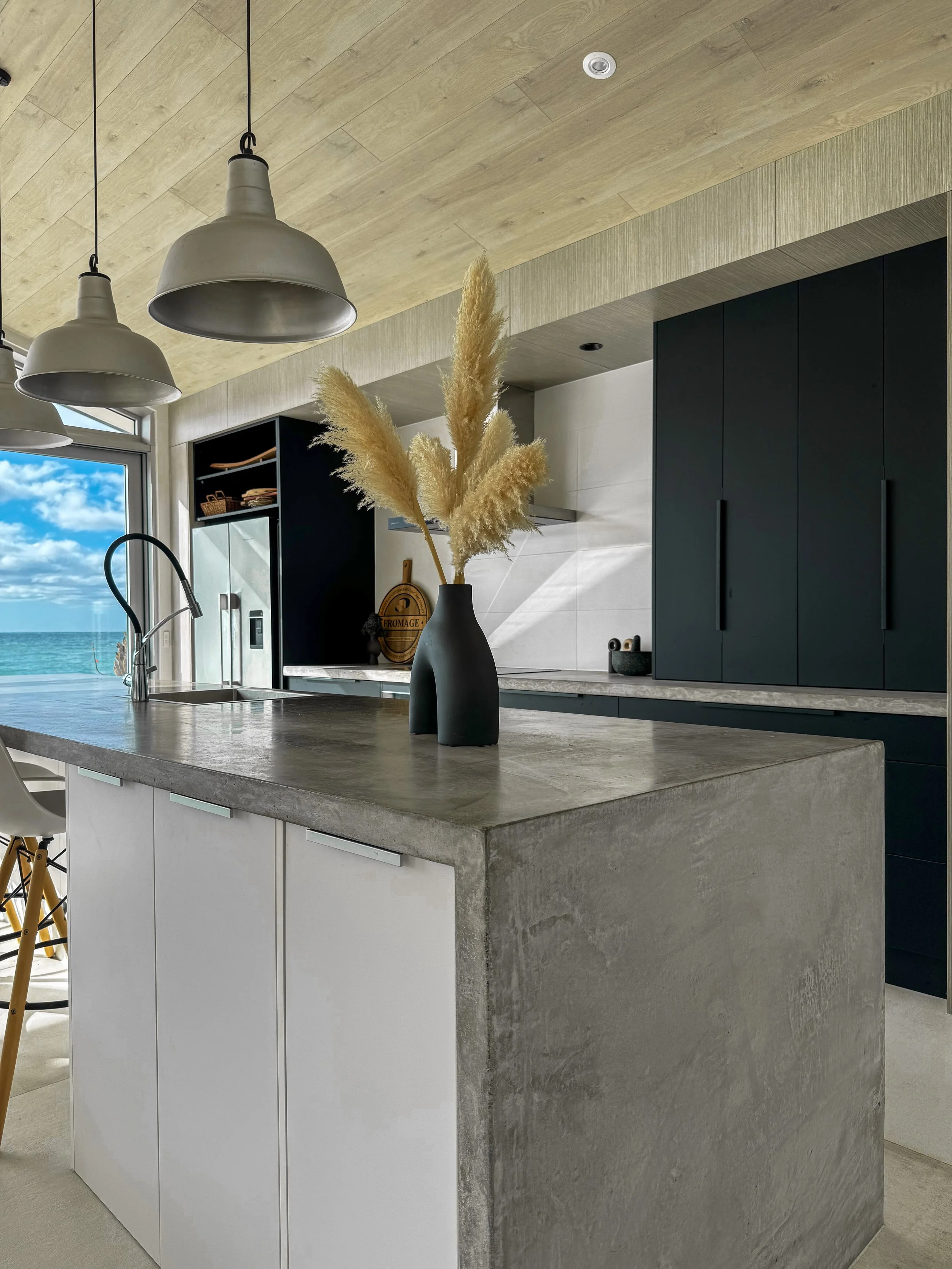 Modern kitchen with a concrete island, black cabinets, a vase with tall dried pampas grass, pendant lights, and a window with an ocean view. Designed by McMahon interior Designer based in Auckland's North Shore.