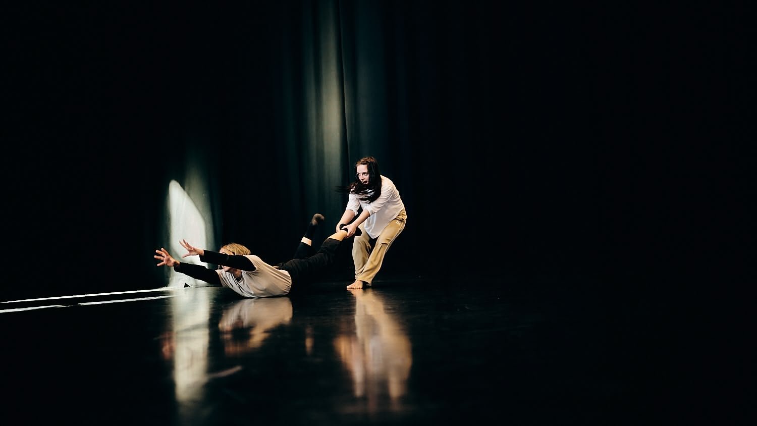 Two people, one on the floor and one standing, on a dark stage with minimal lighting, possibly involved in a dance or theatrical performance.