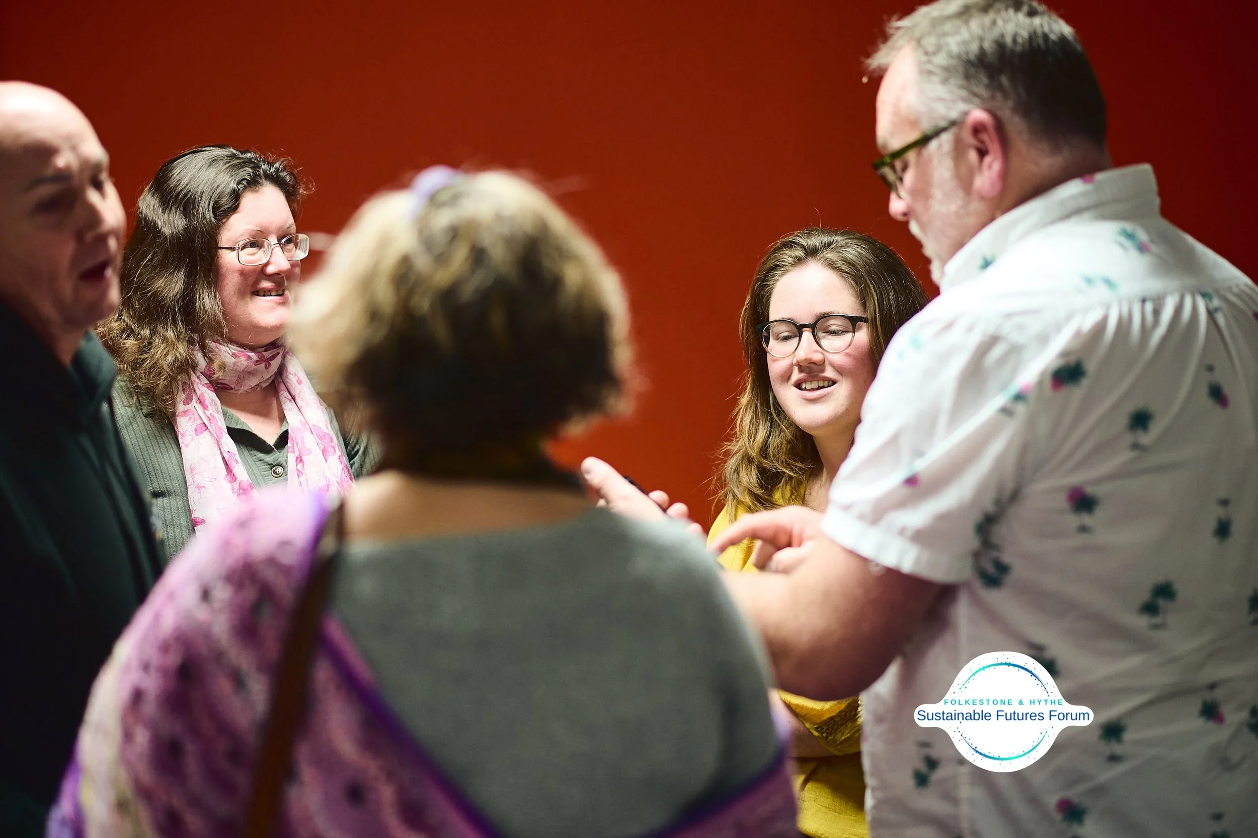 Group of five adults talking and smiling during the Folkeson & Hythe Sustainable Futures Forum, against a red background.