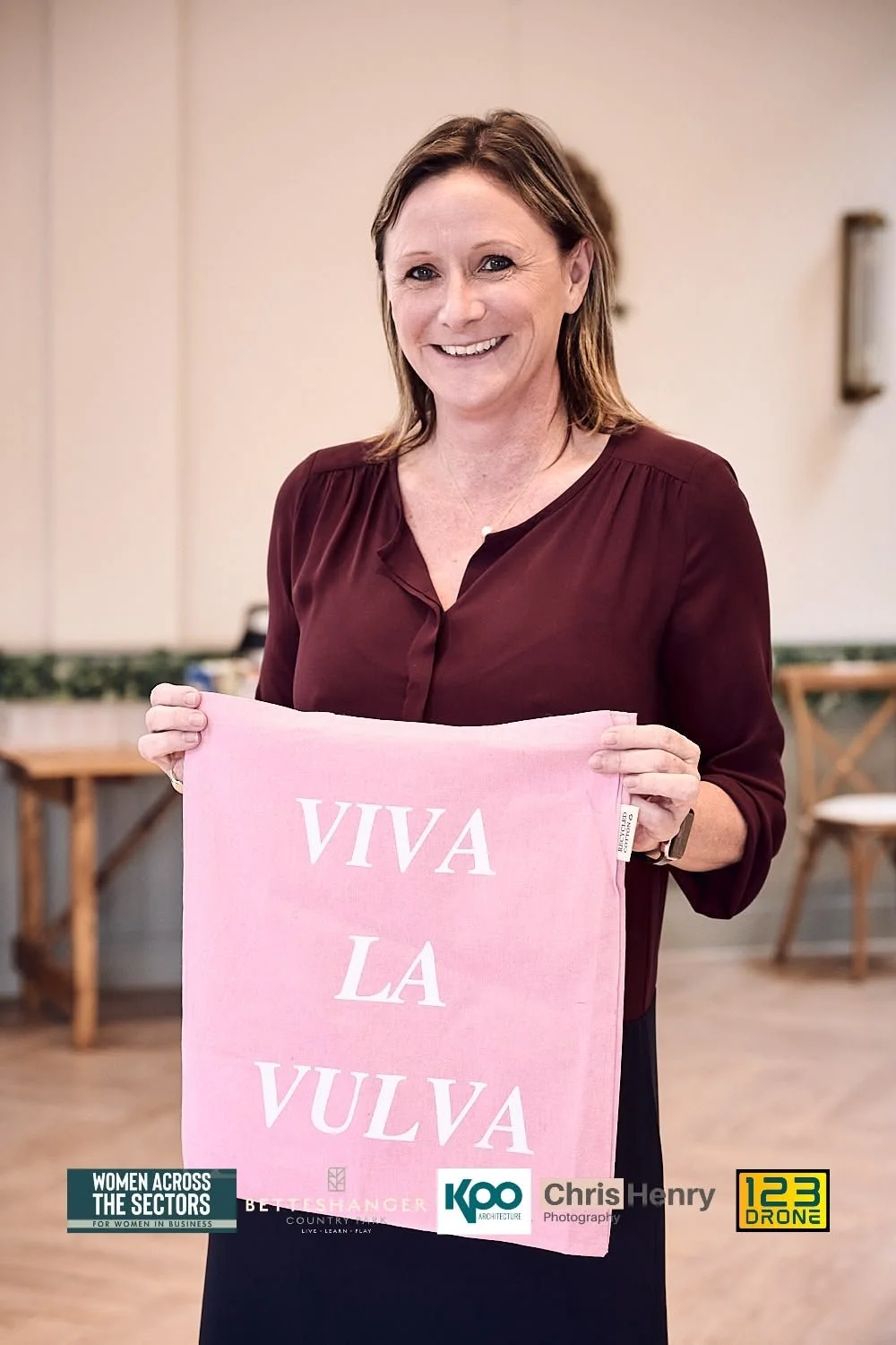 A woman with shoulder-length brown hair, smiling, holding a pink cloth with the words "VIVA LA VULVA" printed on it. She is wearing a dark red blouse and standing indoors with a blurred background of chairs and tables.