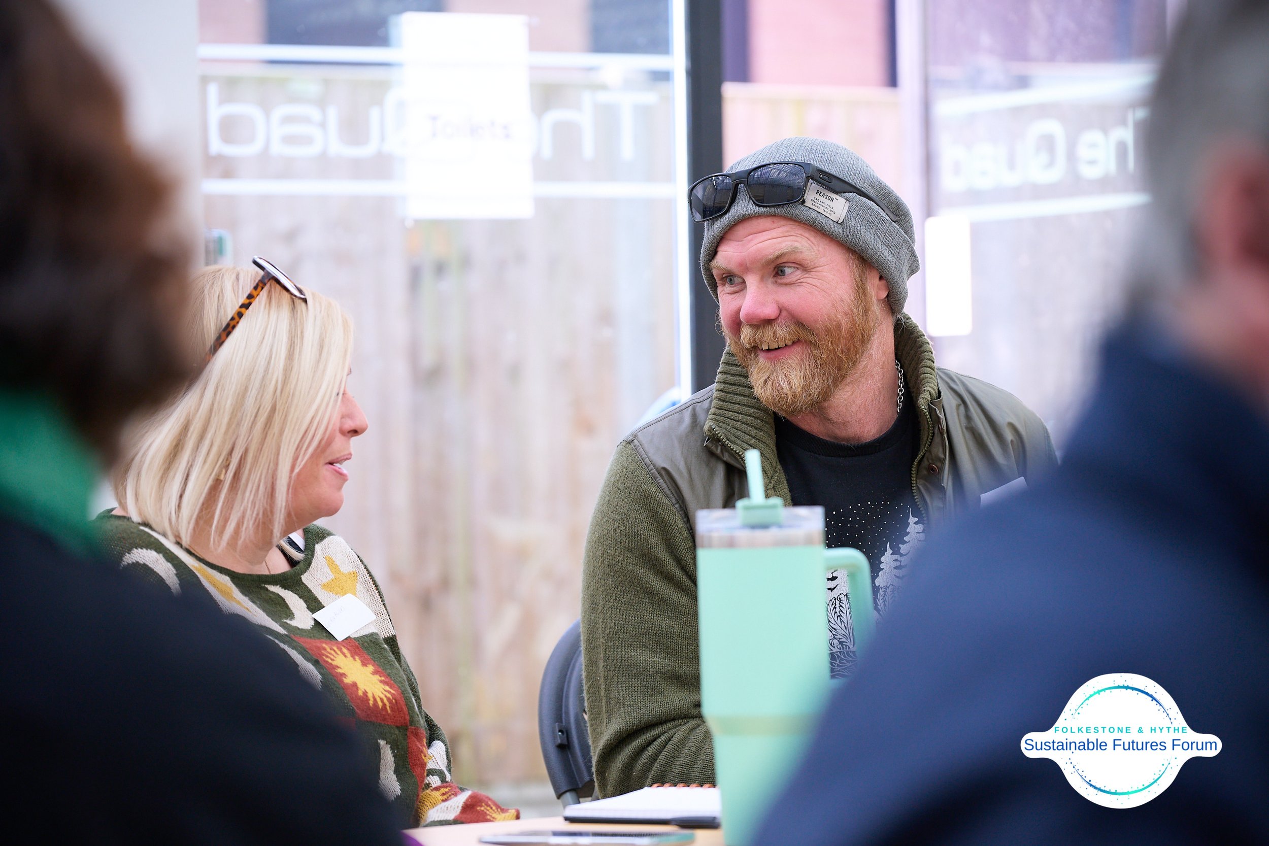 A group of people sitting together at a table indoors, with a man in a gray beanie and sunglasses on his head smiling and talking to a woman with blonde hair wearing glasses and a colorful sweater.