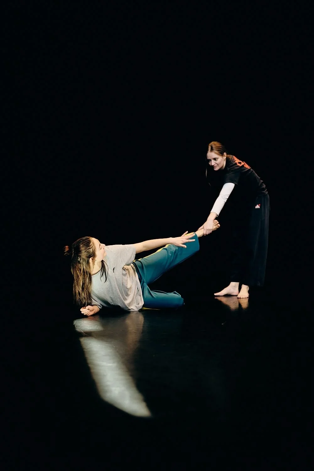 One person is lying on the floor and pulling the leg of another person who is standing. The scene is set against a black background.
