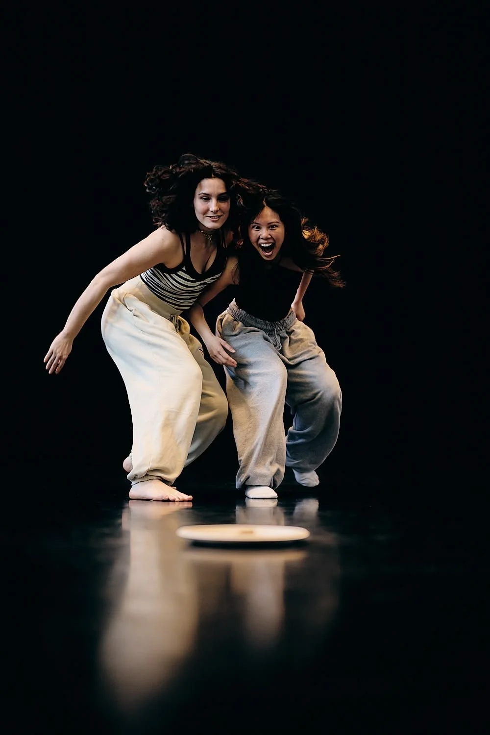 Two women bend down and look at a small object on the floor, with a black background.