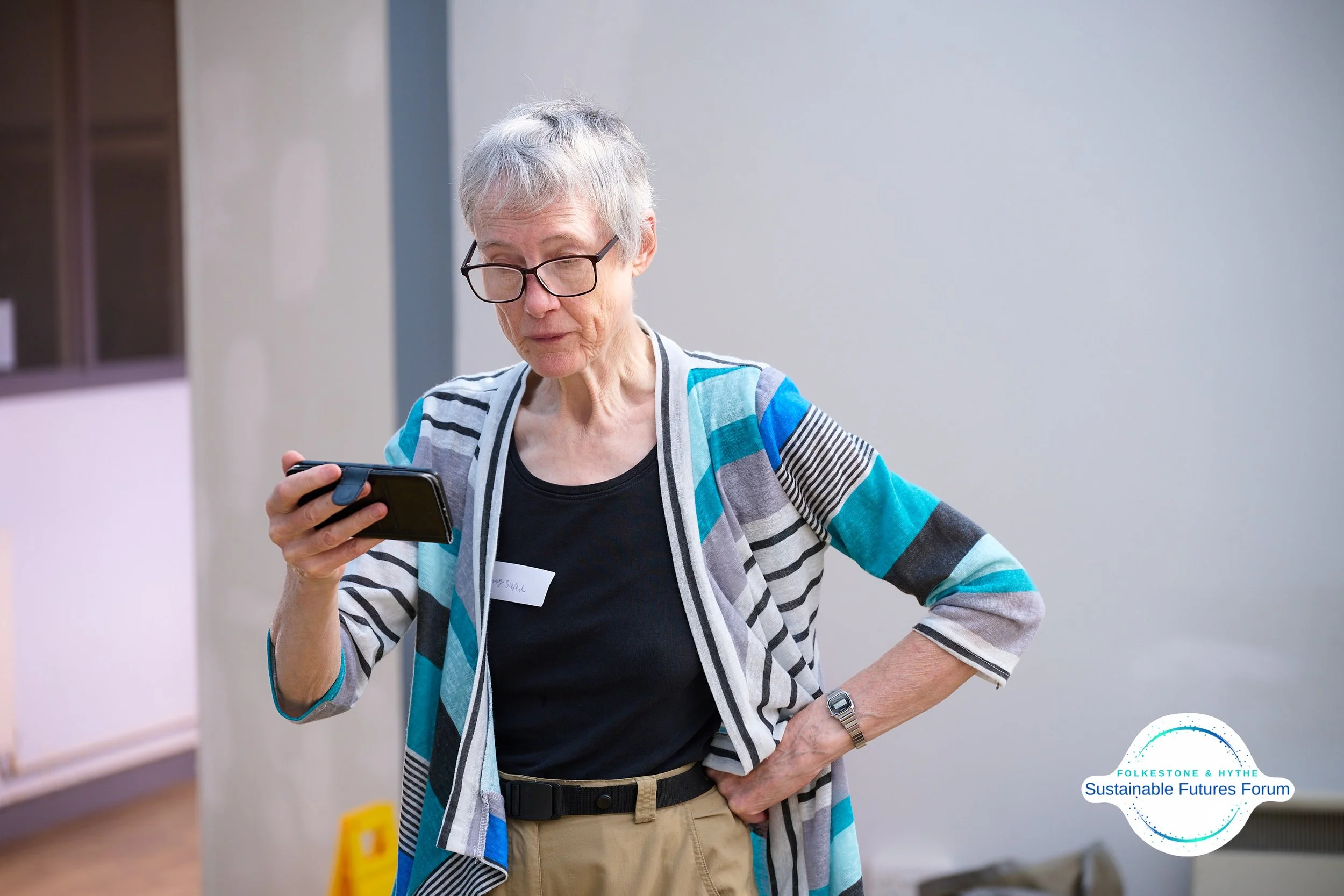An elderly woman with short gray hair, glasses, wearing a colorful striped cardigan over a black shirt, looking at her cellphone at a forum event, with a 'Sustainable Futures Forum' logo in the bottom right corner.