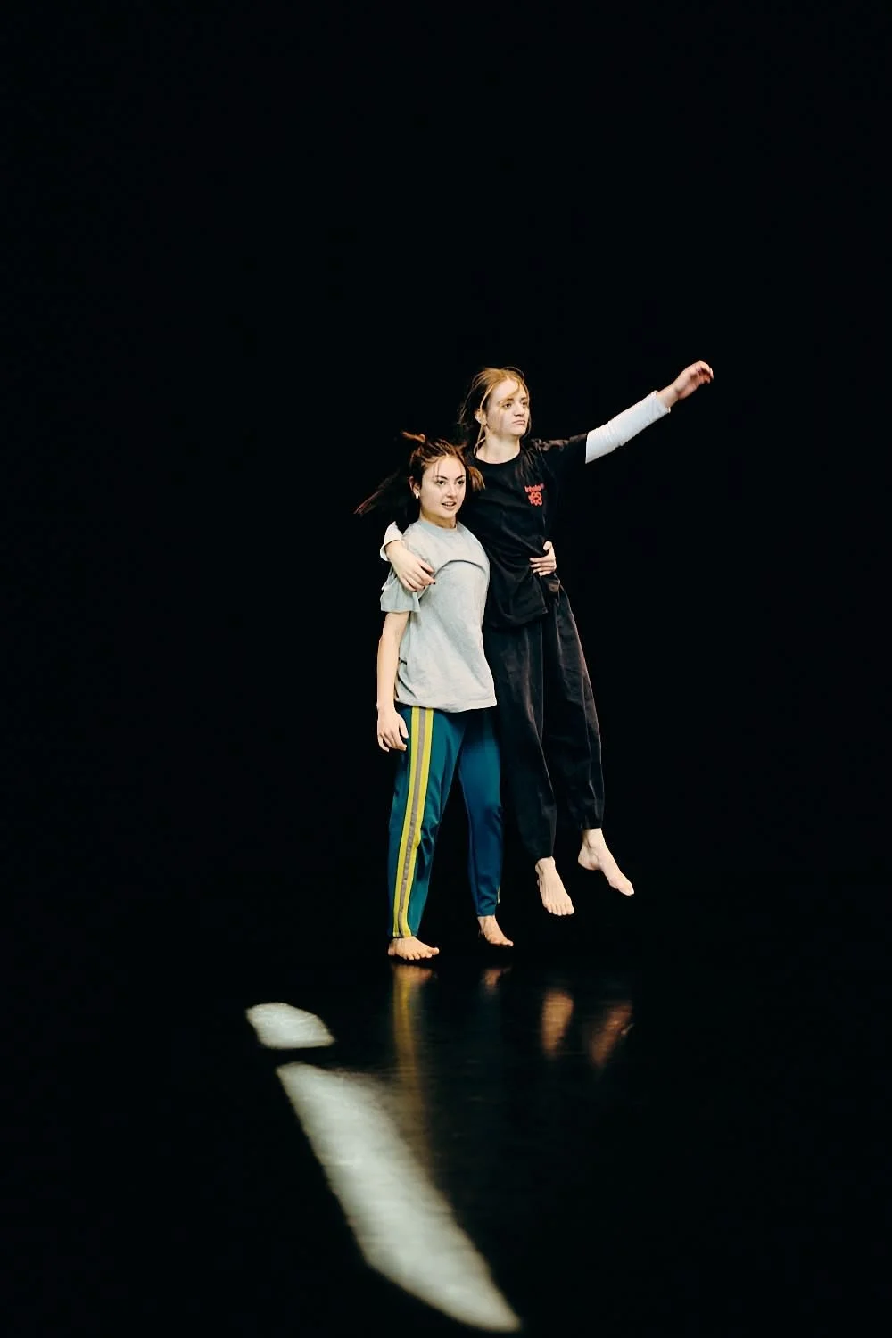 Two young women are dancing or performing on a dark stage. One has her arm around the other's shoulders, and the other is raising her arm. They are barefoot and wearing casual clothes.