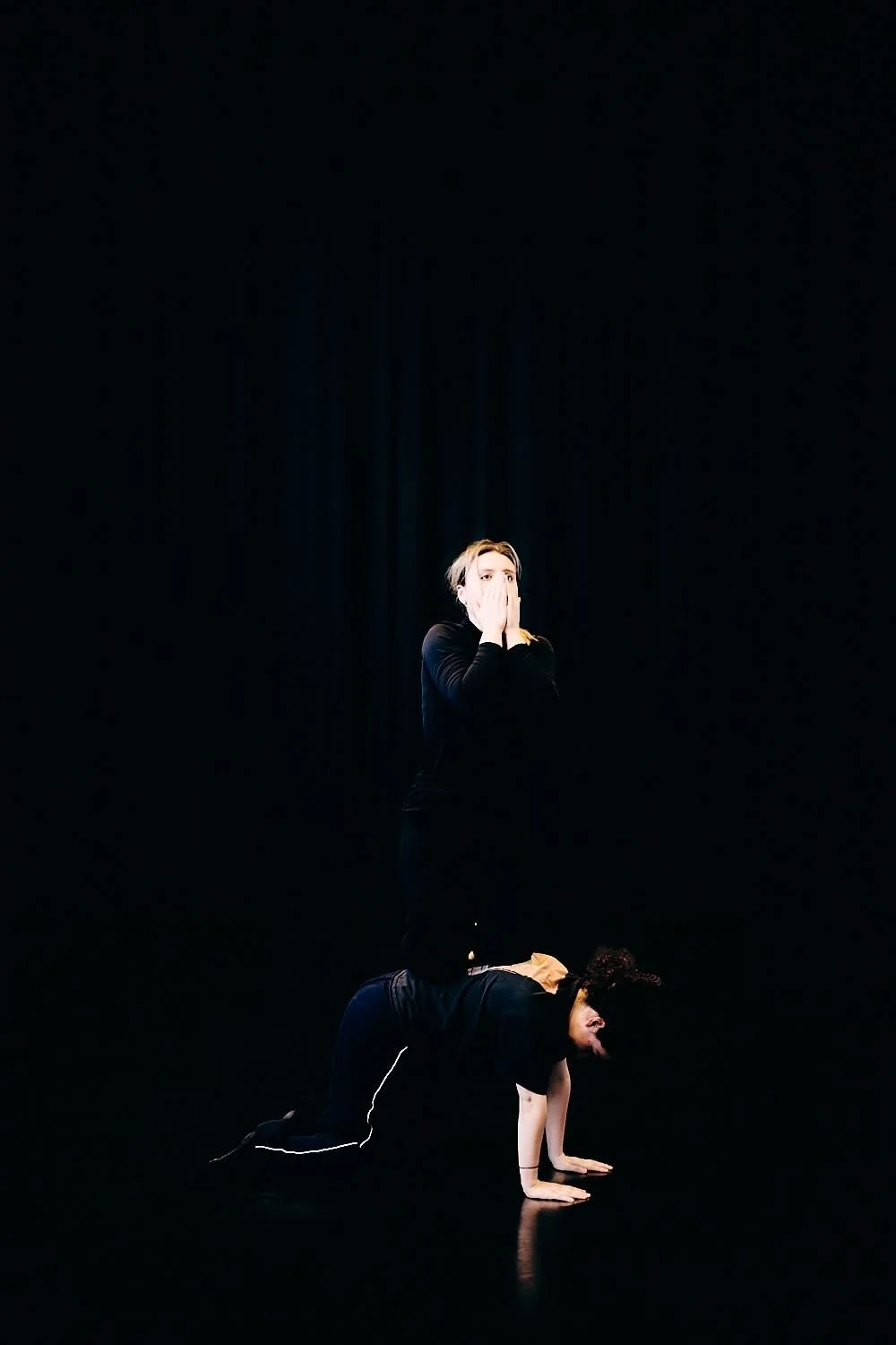 Two women performing an acro yoga pose on stage with a black background. One is in a plank position on the floor, supporting the other woman who is standing on her back with hands covering her mouth.
