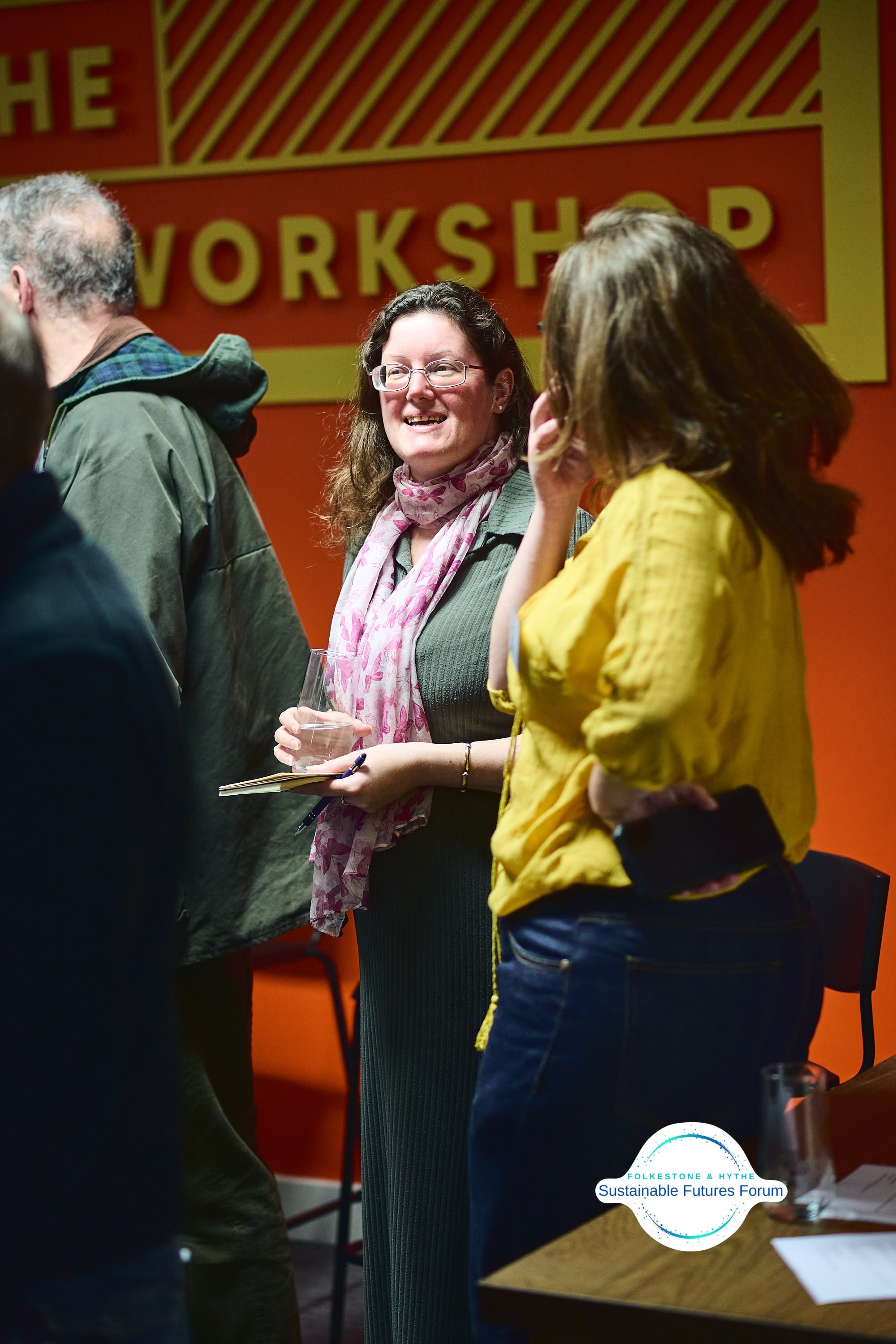 A woman with glasses and curly hair holding a glass of water and a notebook, smiling at a social event with a red and yellow workshop sign in the background.