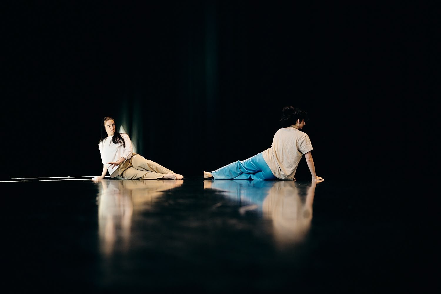 Two performers on a dark stage, one lying on their side and the other on their hands and knees, both facing away from each other.