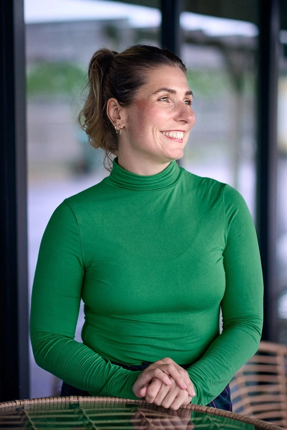 A woman with short, wavy hair, wearing a bright green turtleneck, smiling and standing indoors with a blurred background.
