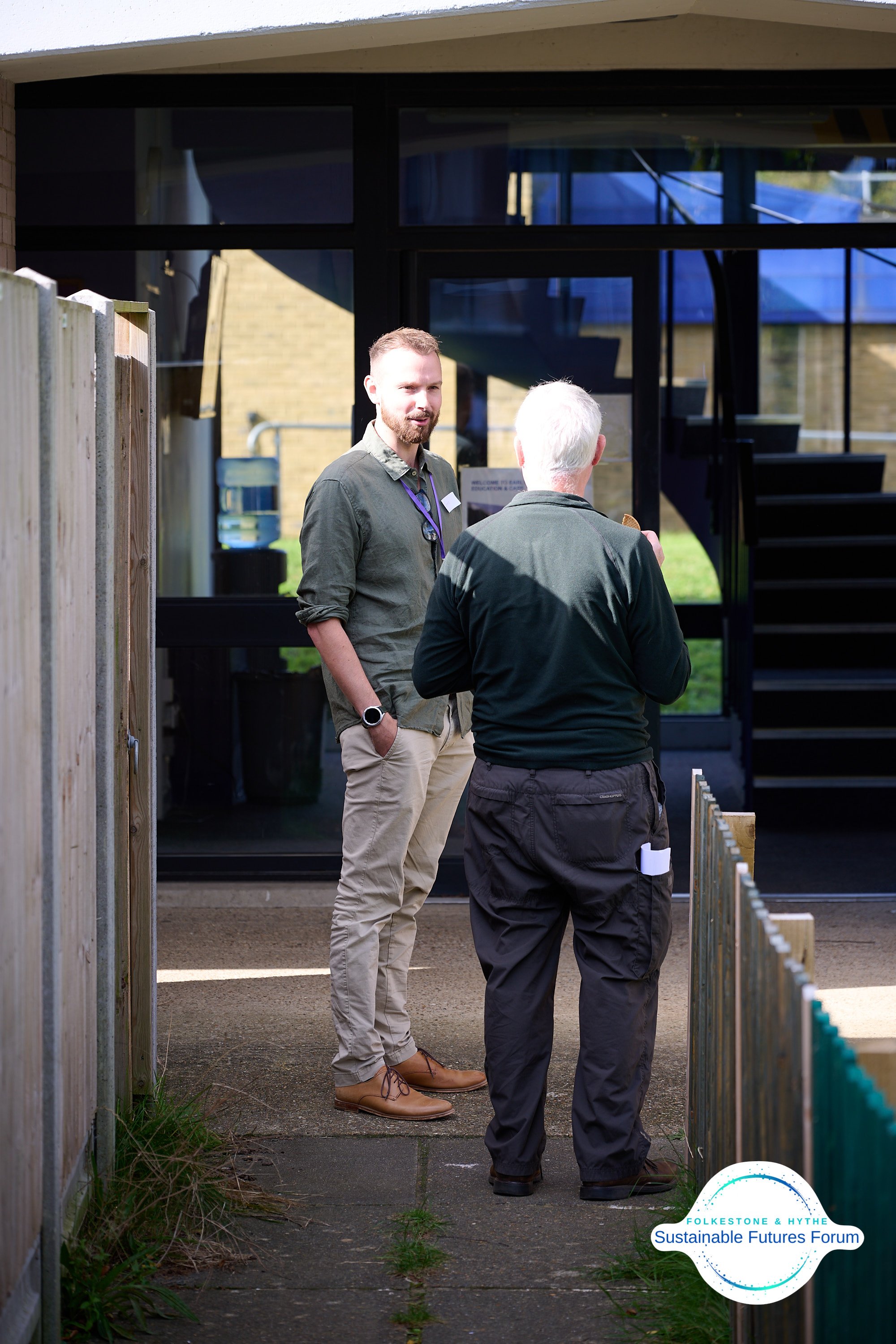 Two men having a conversation outside a building with glass doors. One man has gray hair and wears a black jacket; the other has light brown hair, a beard, and is dressed in a green shirt and beige pants. The image includes the Folkesone & Hythe Sust