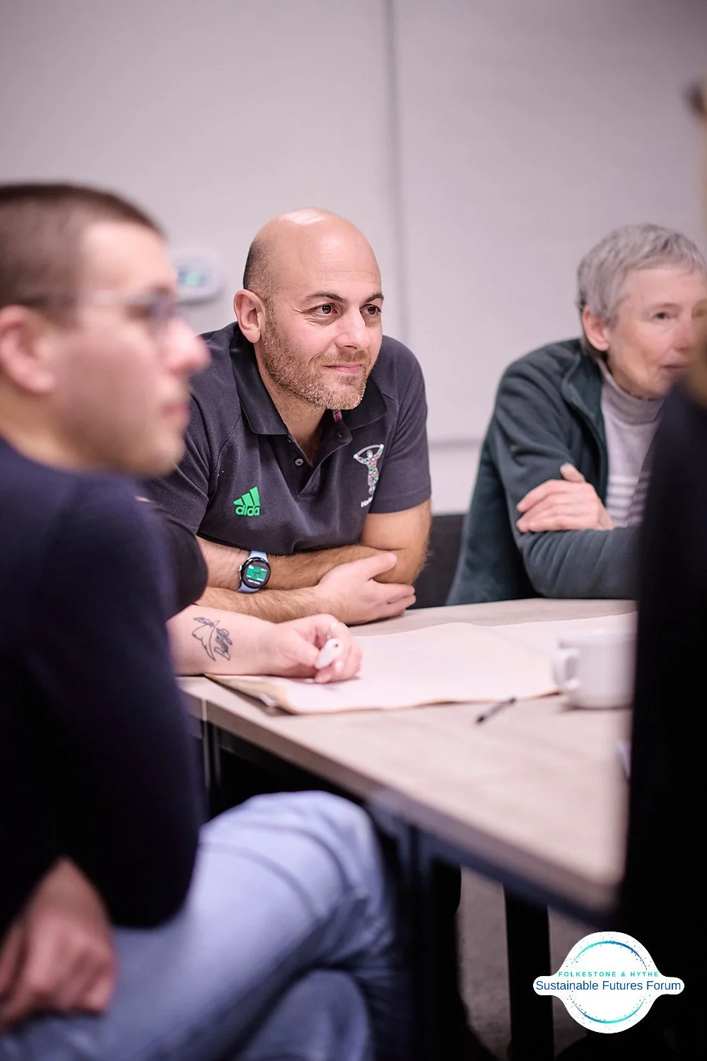 A group of people sitting at a conference table during a meeting, with a focus on a bald man in a black polo shirt with a logo, listening attentively.