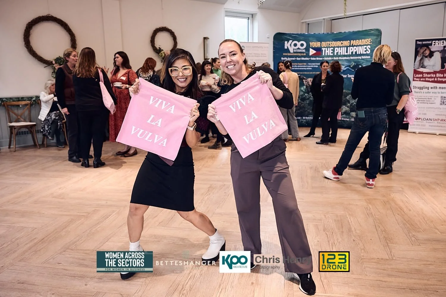 Two women smiling and holding pink signs with white text that reads "VIVA LA VULVA" at a women's business event with other attendees in the background.