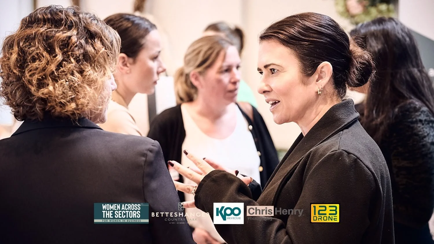 Group of women having a discussion at a professional event, with logos for 'Women Across The Sectors,' 'Bettshanger Country Park,' 'Koo Architecture,' 'Chris Henry Photography,' and '123 Drone' at the bottom.