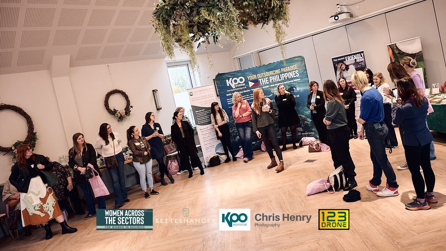 A group of women is standing in a semi-circle, listening to a speaker at an indoor event with arranged decorations, banners, and informational displays.