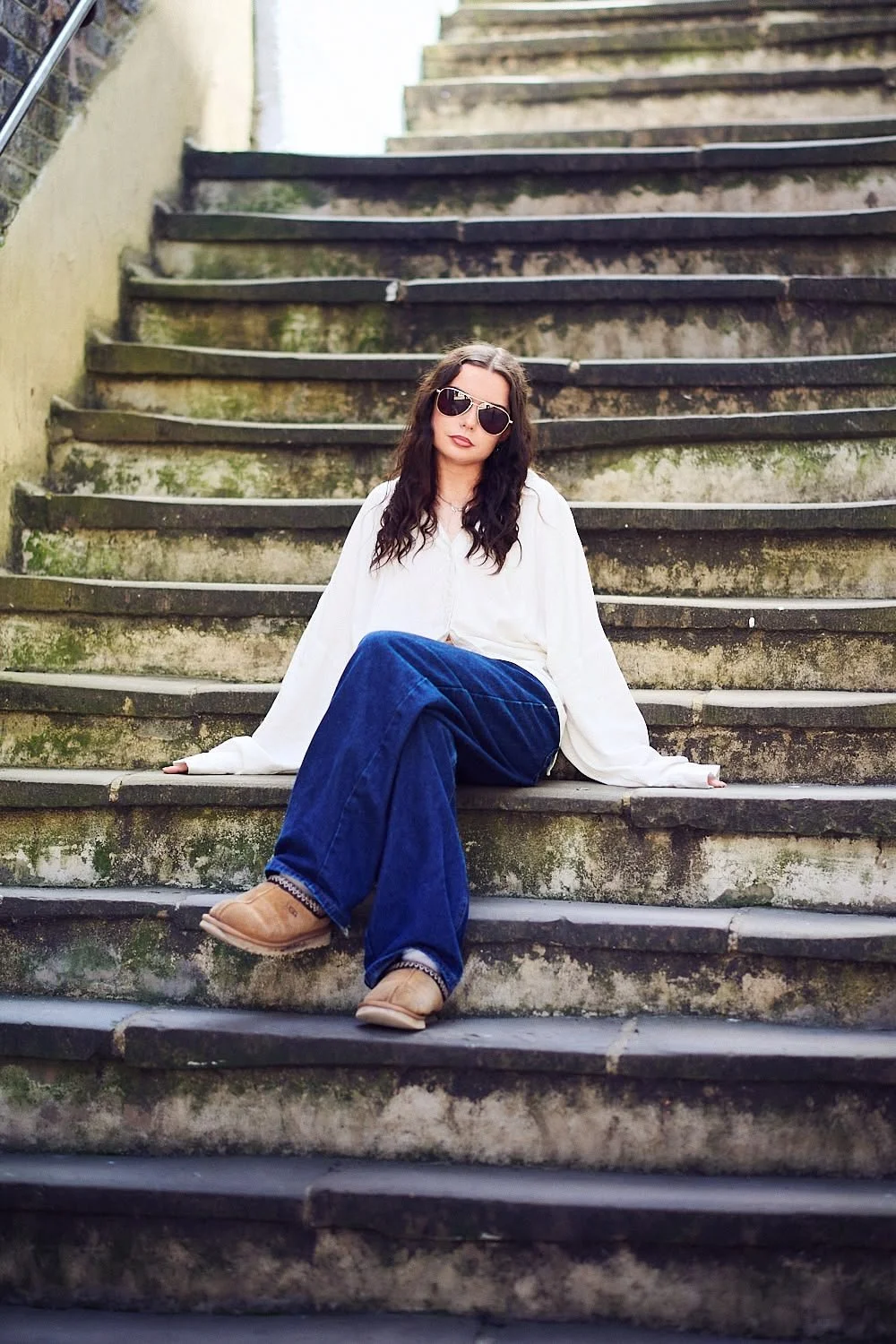 A woman with long dark hair, sunglasses, a white blouse, blue wide-leg jeans, and tan boots, sitting on a mossy, weathered outdoor staircase with a relaxed pose.