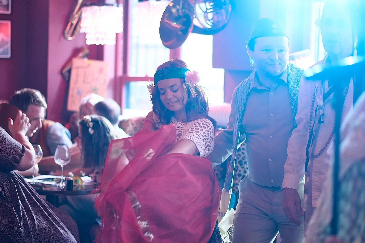 People celebrating at a party in a cozy, colorful room with red walls, decorated with musical instruments and neon lights. A woman wearing a flower crown and a crochet top stands next to a man in traditional German attire, smiling amid a festive atmo
