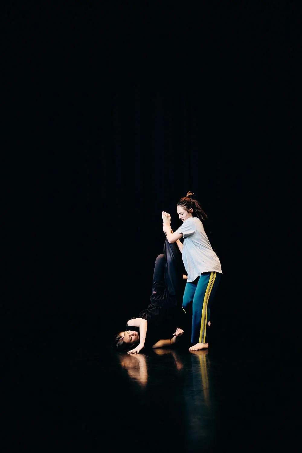 Two women practicing Brazilian Jiu-Jitsu on a dark, minimalistic stage. One woman is on the ground holding the other woman in a submission, while the standing woman is preparing to maneuver her.