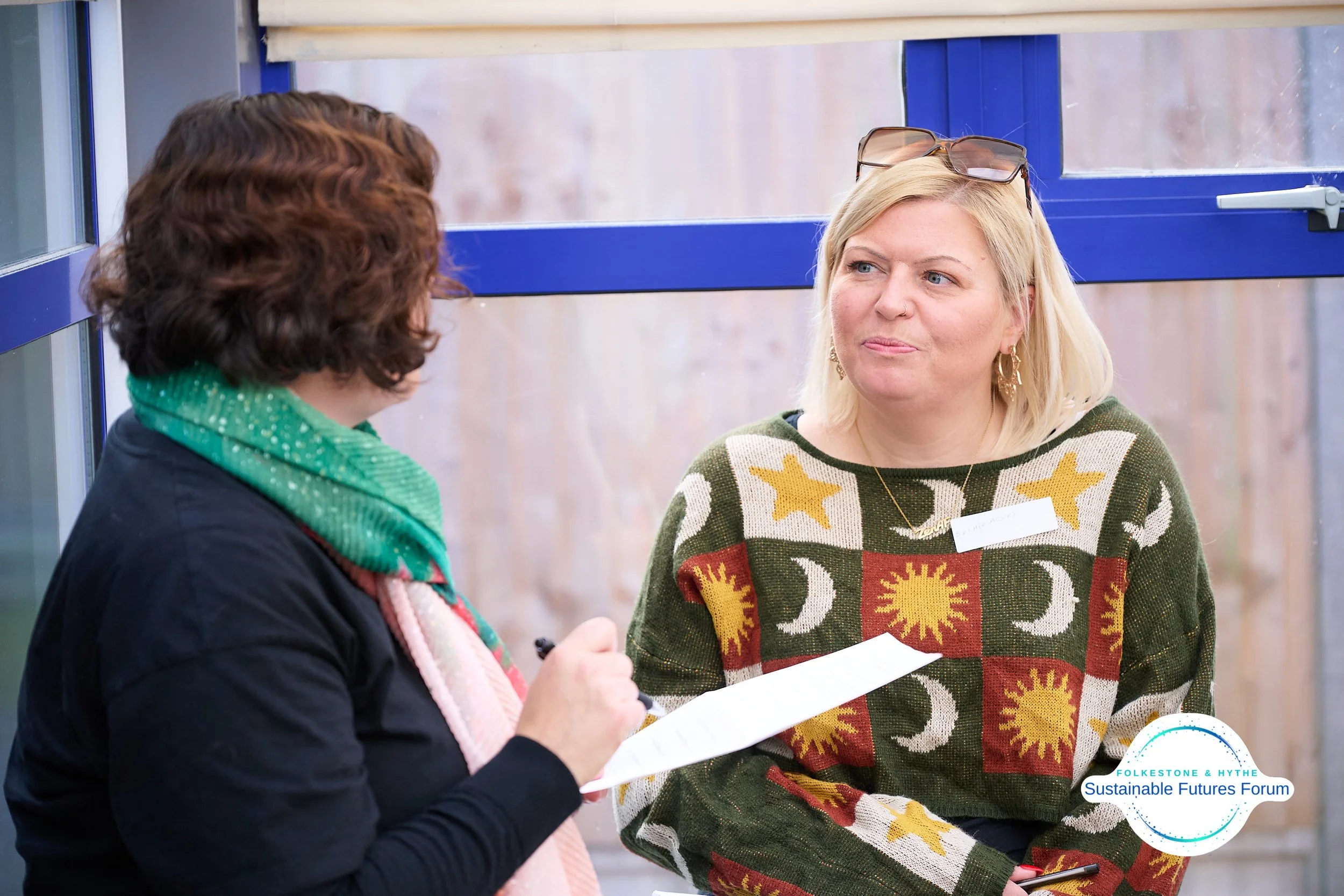 Two women having a conversation indoors, one with curly brown hair and green scarf, the other with blonde hair, glasses on her head, wearing a colorful sweater with sun, moon, and star patterns, at a sustainable futures forum.
