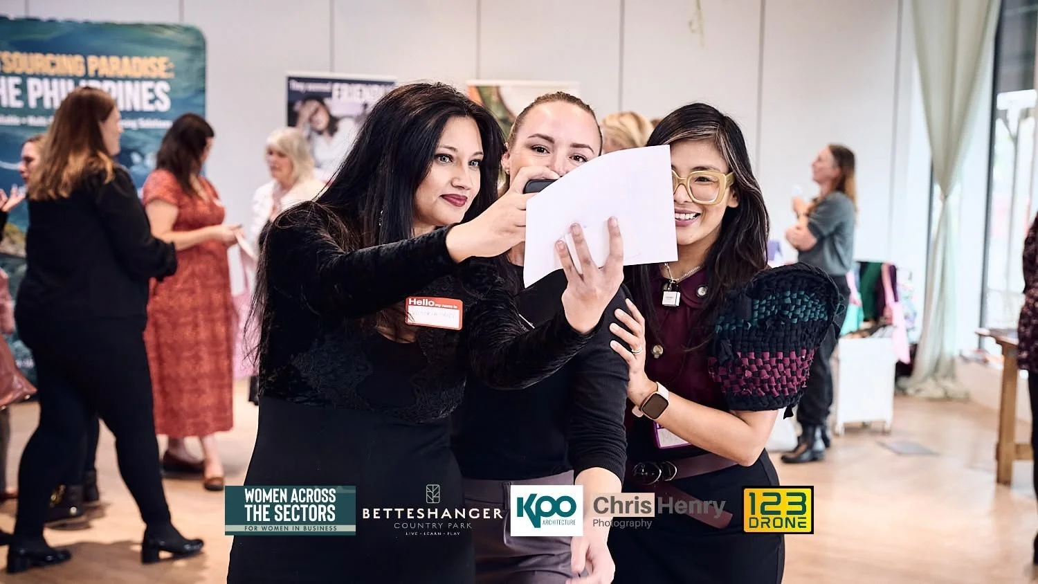 Three women taking a selfie at a networking event, with other women in the background.