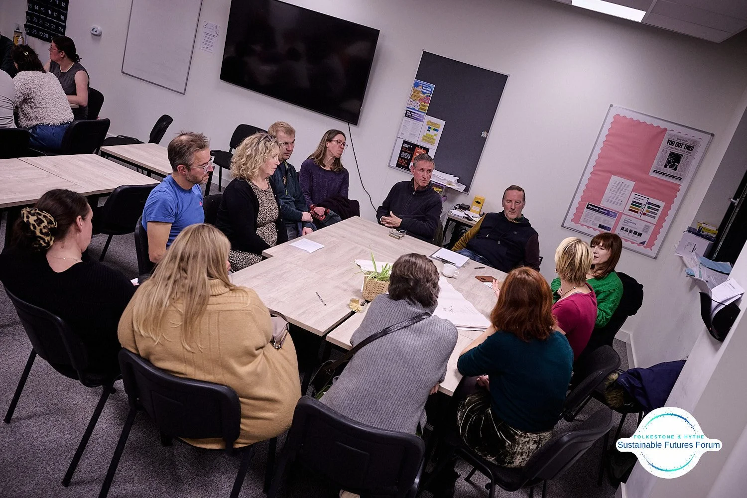 A group of people seated around a rectangular table in a conference room, engaged in a discussion, with a large TV screen and posters on the walls in the background.