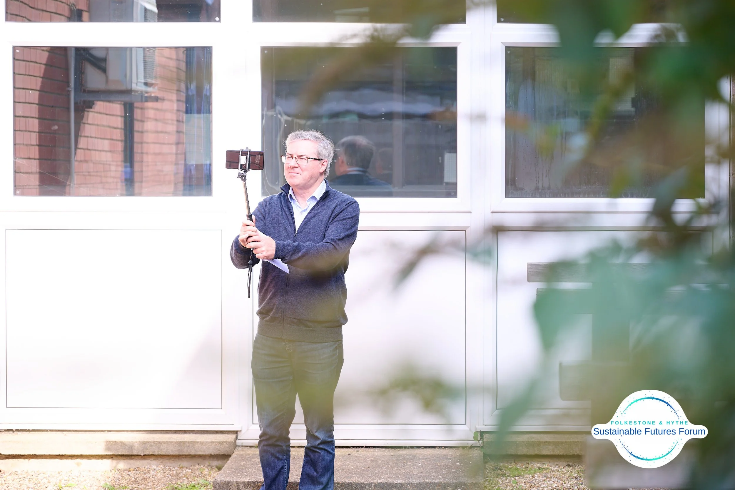 A man with gray hair and glasses standing outside, holding a selfie stick with a smartphone, in front of a white building with large windows. There is a logo in the bottom right corner that reads 'Folkes tone & Hythe Sustainable Futures Forum'.