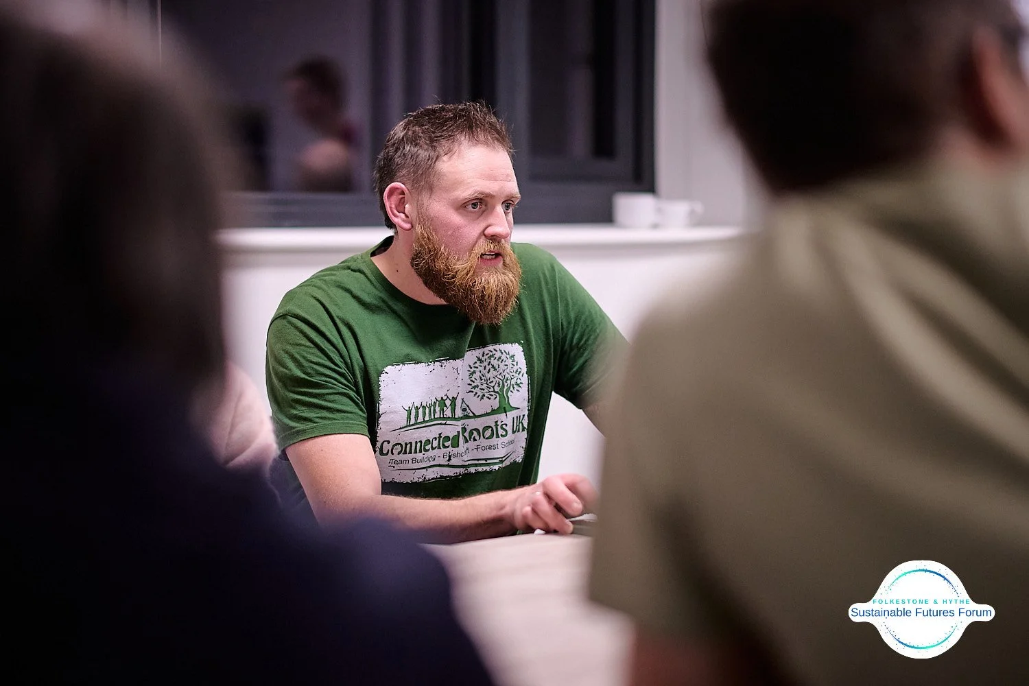 Man with a beard wearing a green t-shirt with a logo and text, sitting at a table in a meeting or discussion, with several people around him in an indoor setting.