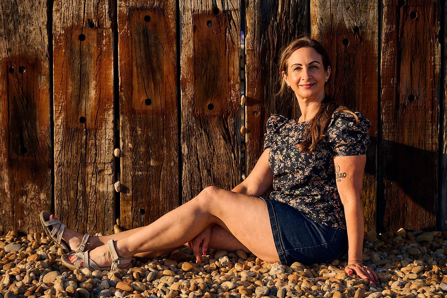 Woman with brown hair sitting on pebbles against a wooden fence, wearing a floral top, denim shorts, and sandals, smiling at the camera.